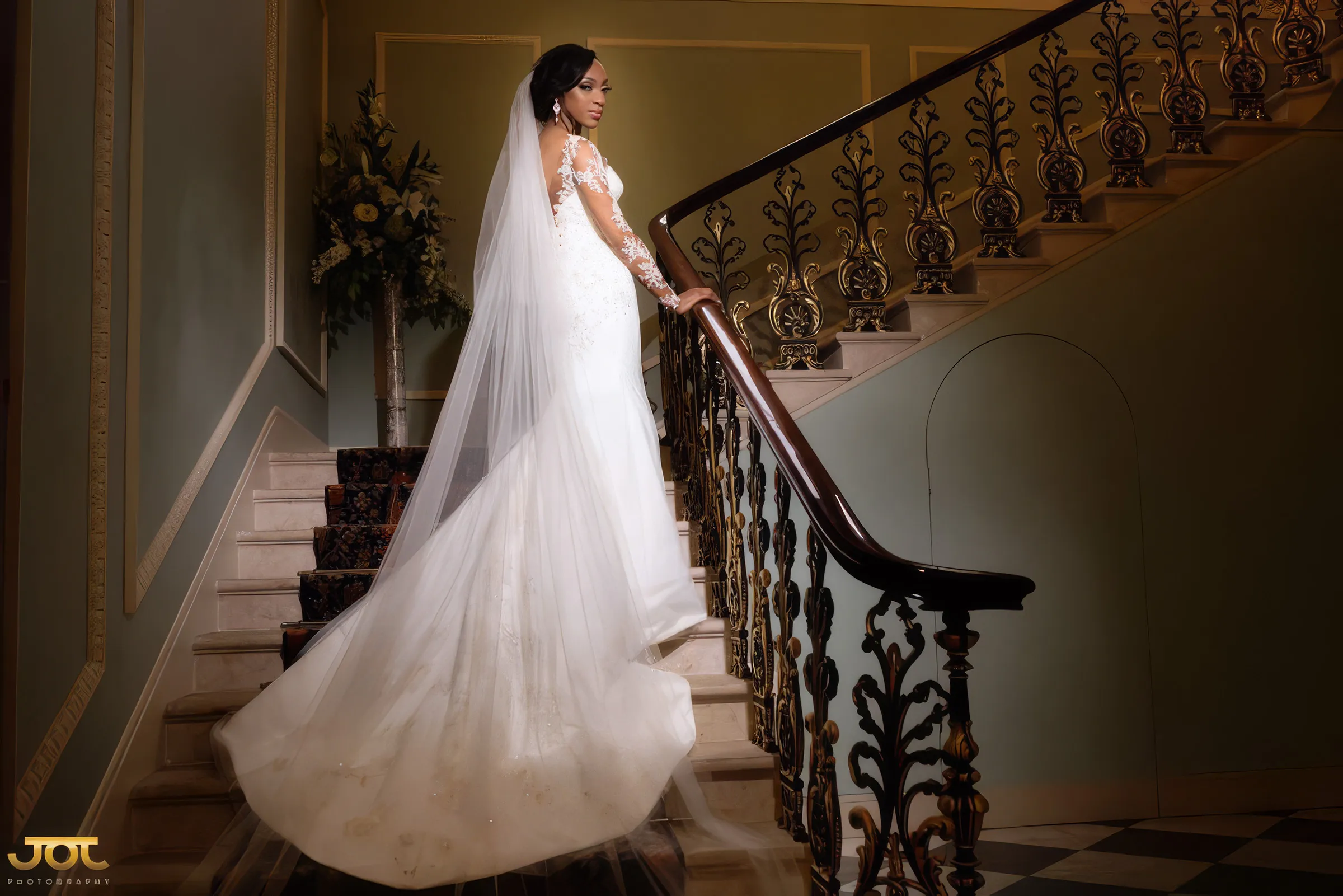 Cherelle in London, looking back from a grand staircase with an ornate wrought-iron balustrade. Her bespoke Kosibah couture gown has long sheer lace sleeves and a flared skirt, with a long tulle veil cascading down the steps.