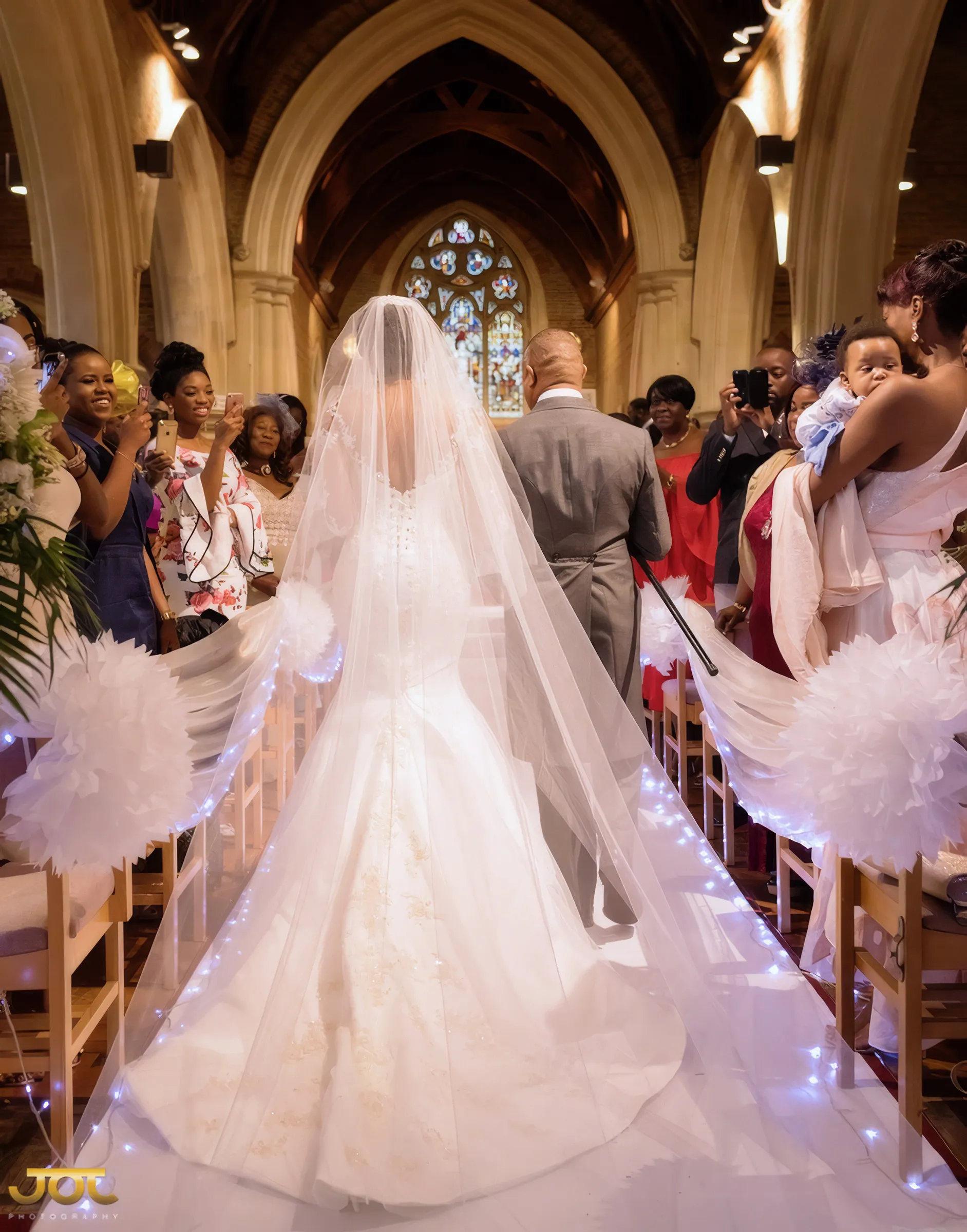 Cherelle seen from behind as she walks down the church aisle with a man in a grey morning suit, her long tulle veil and the dramatic flared train of her gown spreading over the fairy-lit carpet as guests look on.