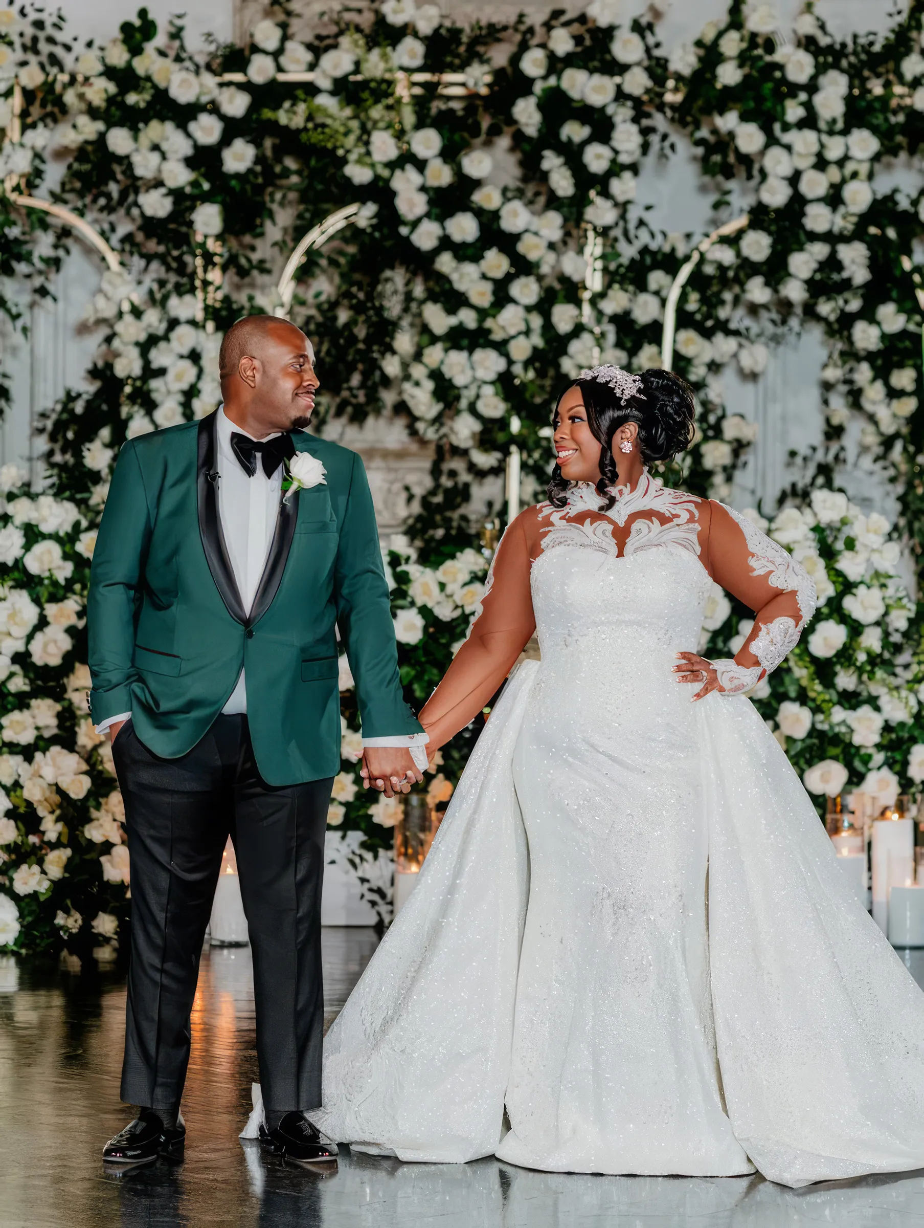 Cathy holds hands with her husband, in an emerald tuxedo, before a wall of white flowers. Her figure-hugging gown has an intricate lace-appliquéd illusion neckline and long sheer sleeves, with the detachable train falling from the waist.