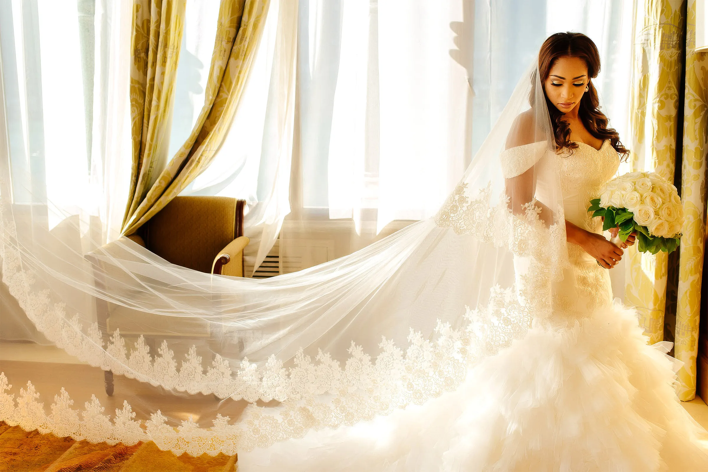 Layal looks down in a sunlit window, her lace-edged veil flowing across the room. Her couture gown has an off-the-shoulder sweetheart bodice and a cascading ruffled tulle skirt.