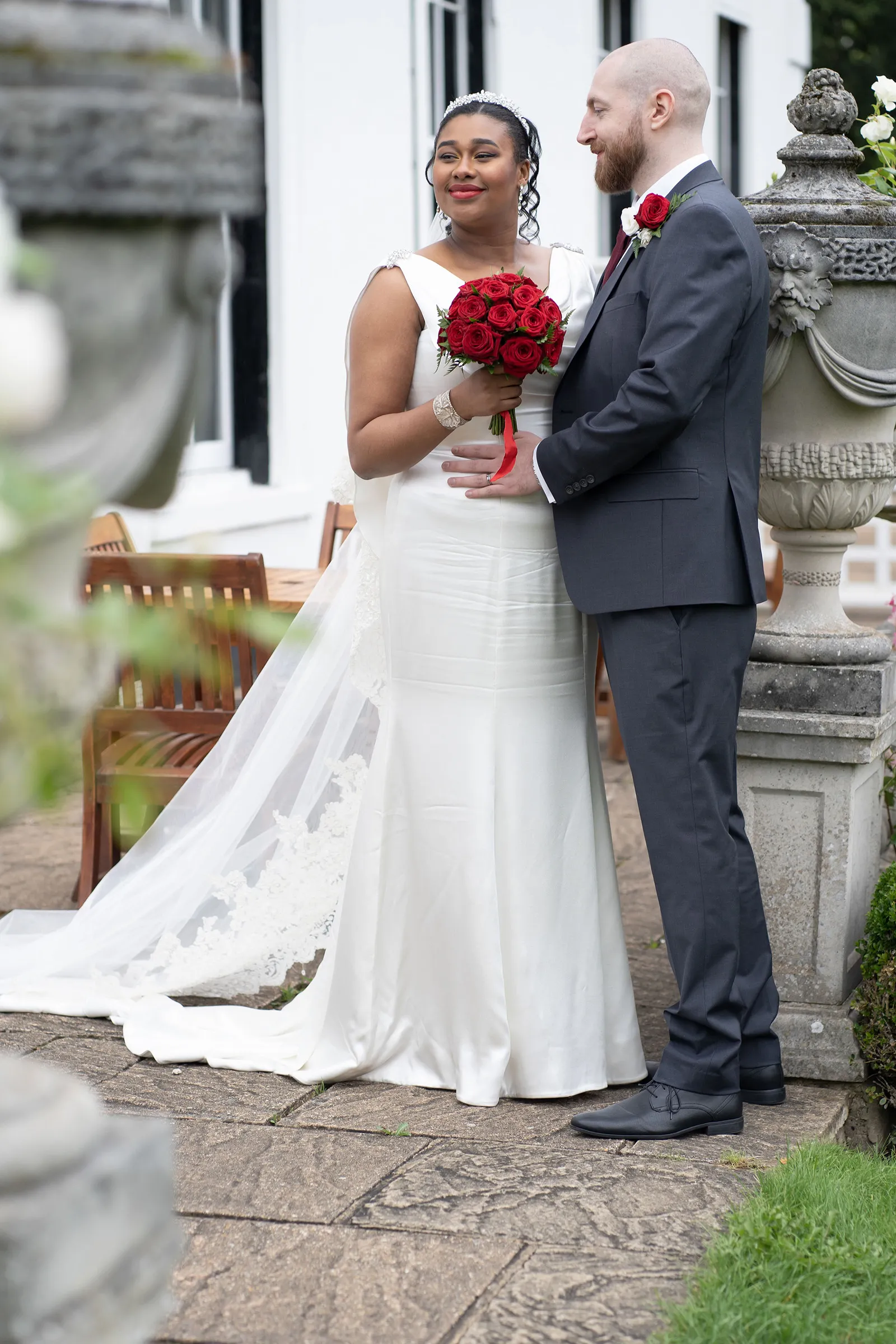 Lola holds a bouquet of red roses beside her husband in a garden. Her sculpted ivory bridal gown has jewelled shoulder accents and a flared hem, with a lace-edged veil trailing behind.