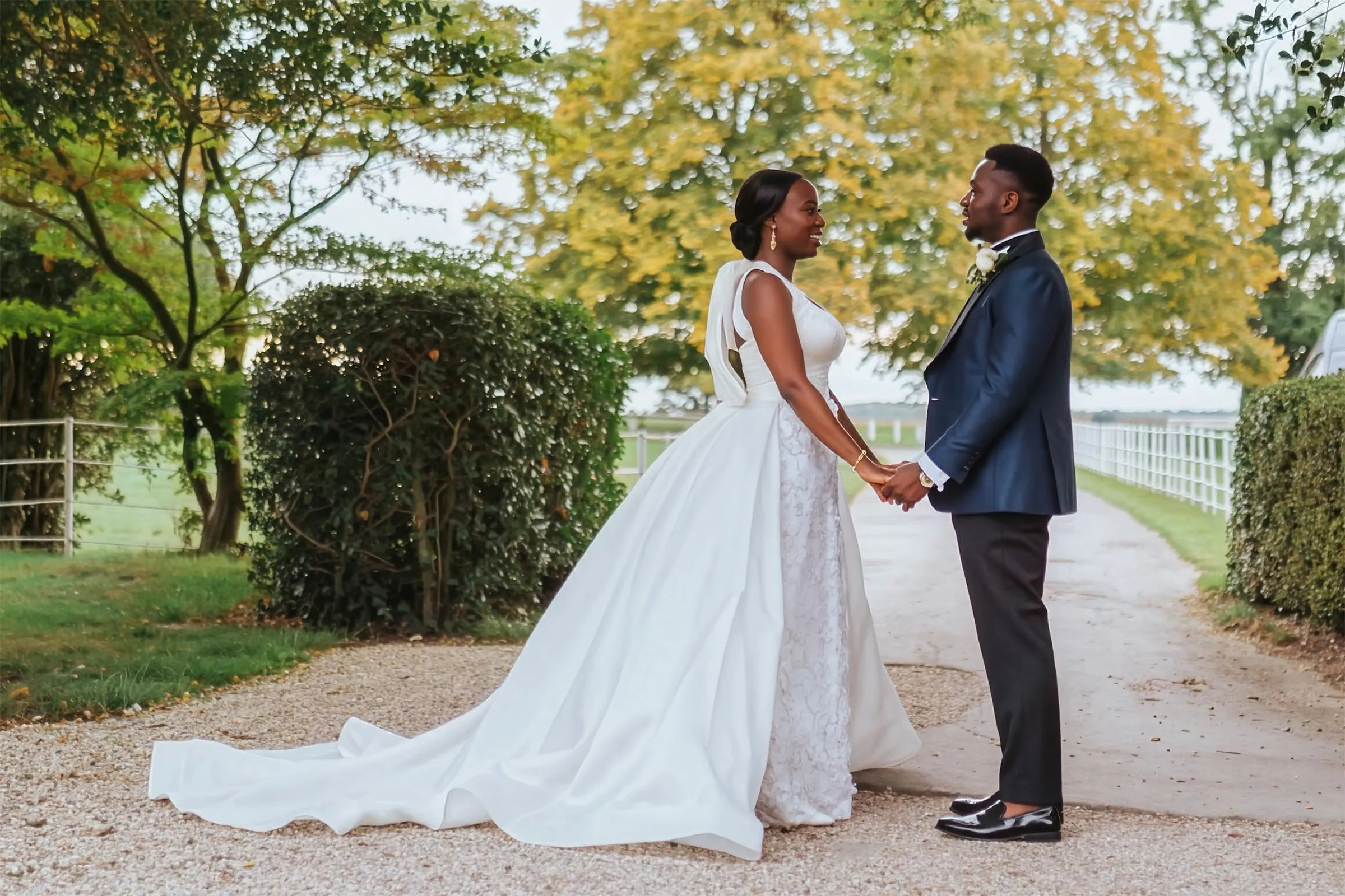 Ife and her husband hold hands on a gravel drive at Barnston Lodge, Essex. She wears a halter-neck bridal gown with a floral-appliquéd skirt and a detachable overskirt with a sweeping train.