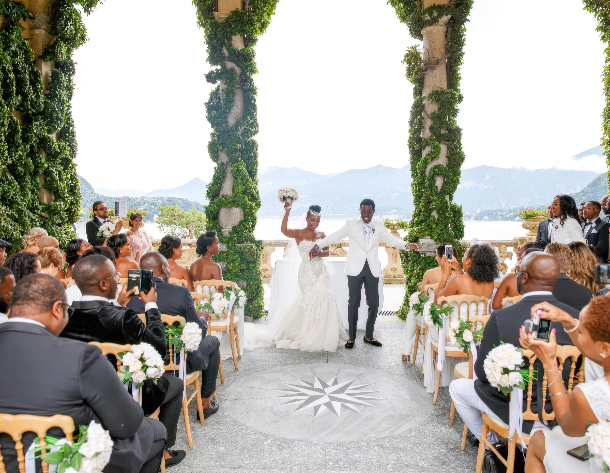 Denise, Lake Como, Italy, holds her husband's hand and smiles, in a strapless sweetheart bridal gown with a soft peach-toned lace bodice and three-dimensional floral appliqués, a jewelled headpiece and a veil.
