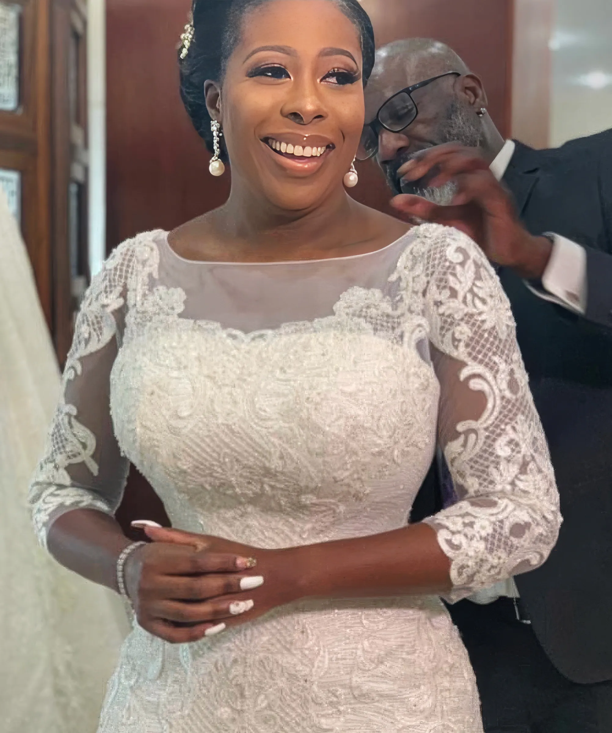 Abies smiles as she gets ready in Lagos, a man in a dark suit helping at the back of her neck. Exquisite lace appliqués cover the illusion neckline, three-quarter-length sleeves and fitted bodice of her figure-hugging gown.