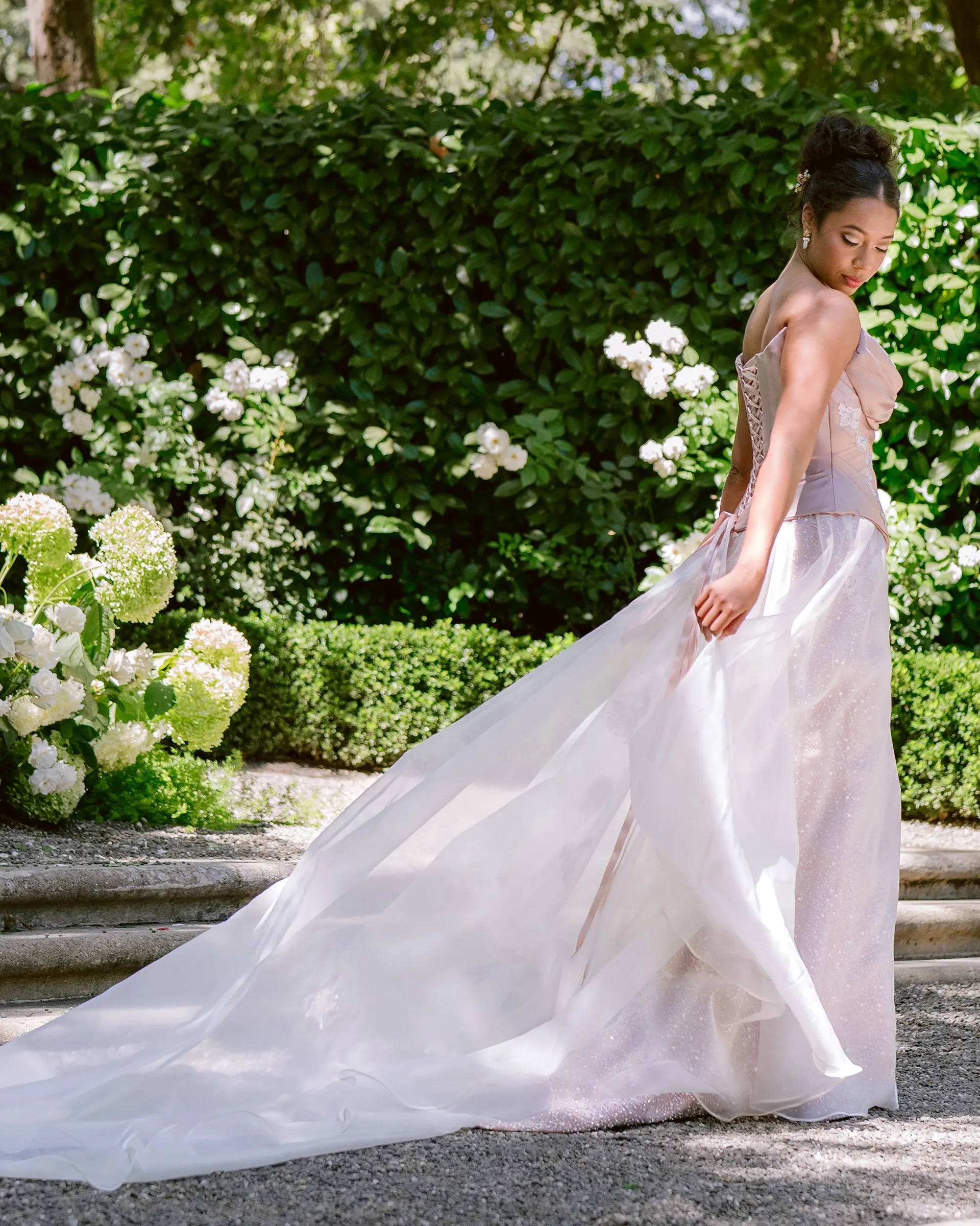 Mist from behind, looking over her shoulder among white hydrangeas: the laced corset back and the sheer organza overskirt sweeping into a long train across the gravel.