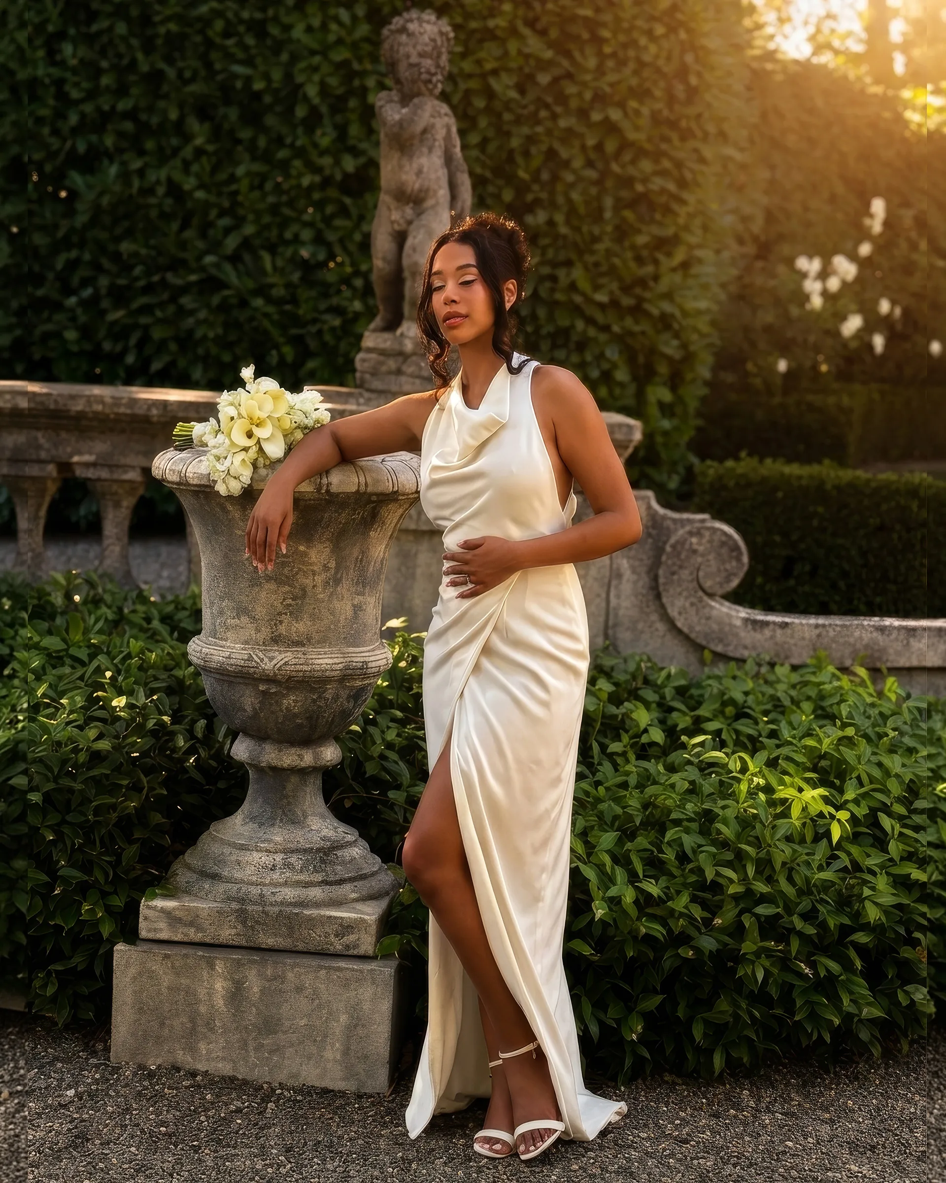 Pearl leaning on a stone urn of white flowers beneath a statue: the cowl neck falls softly at the bust and the wrap skirt drapes from a high slit to the ground.