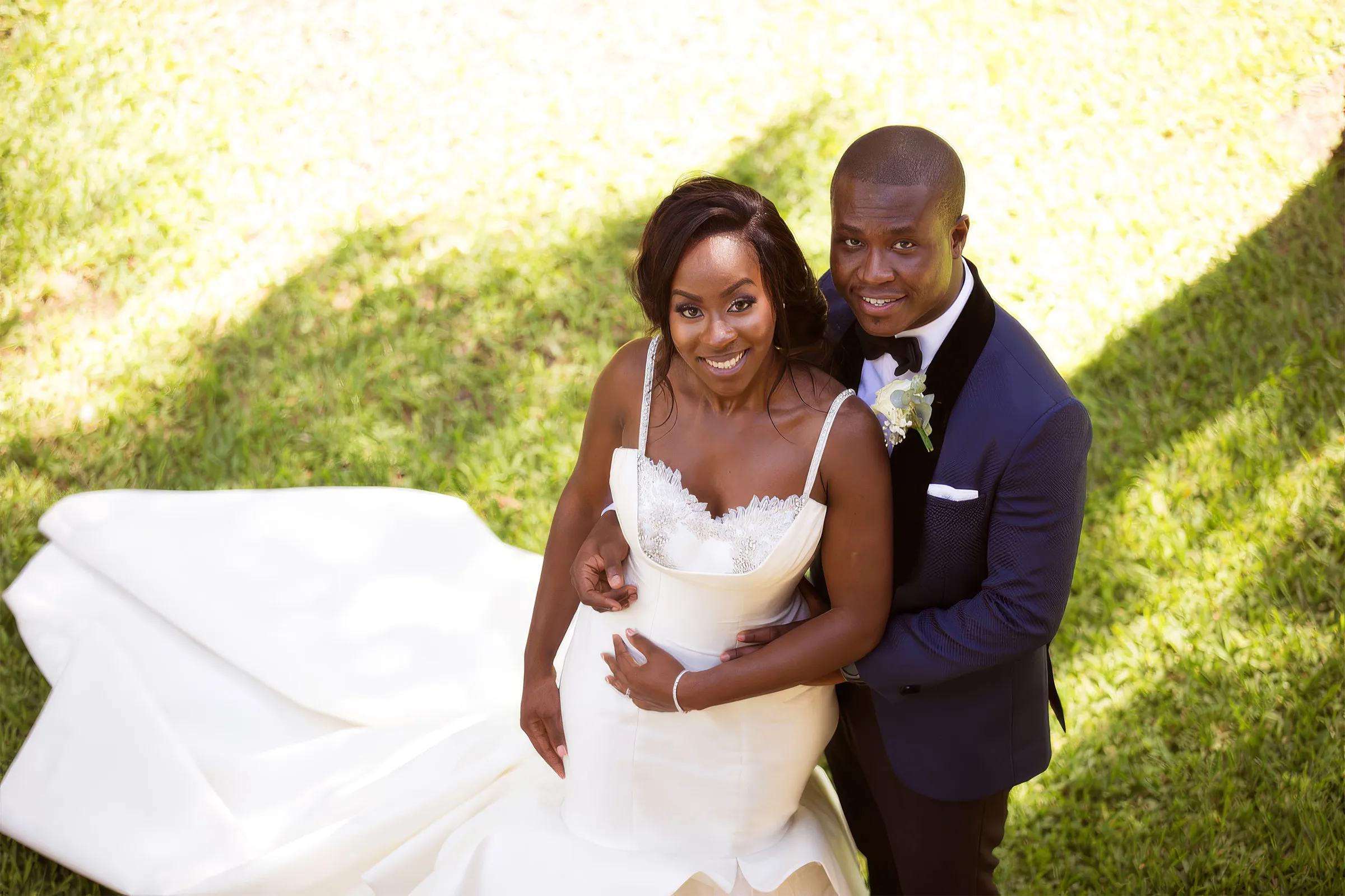 Nana Ama, in Accra, Ghana, smiles up at the camera in her husband's arms on a sunlit lawn. Her gown has a softly draped cowl neckline edged with embellishment and thin jewelled straps, its long train spread across the grass.