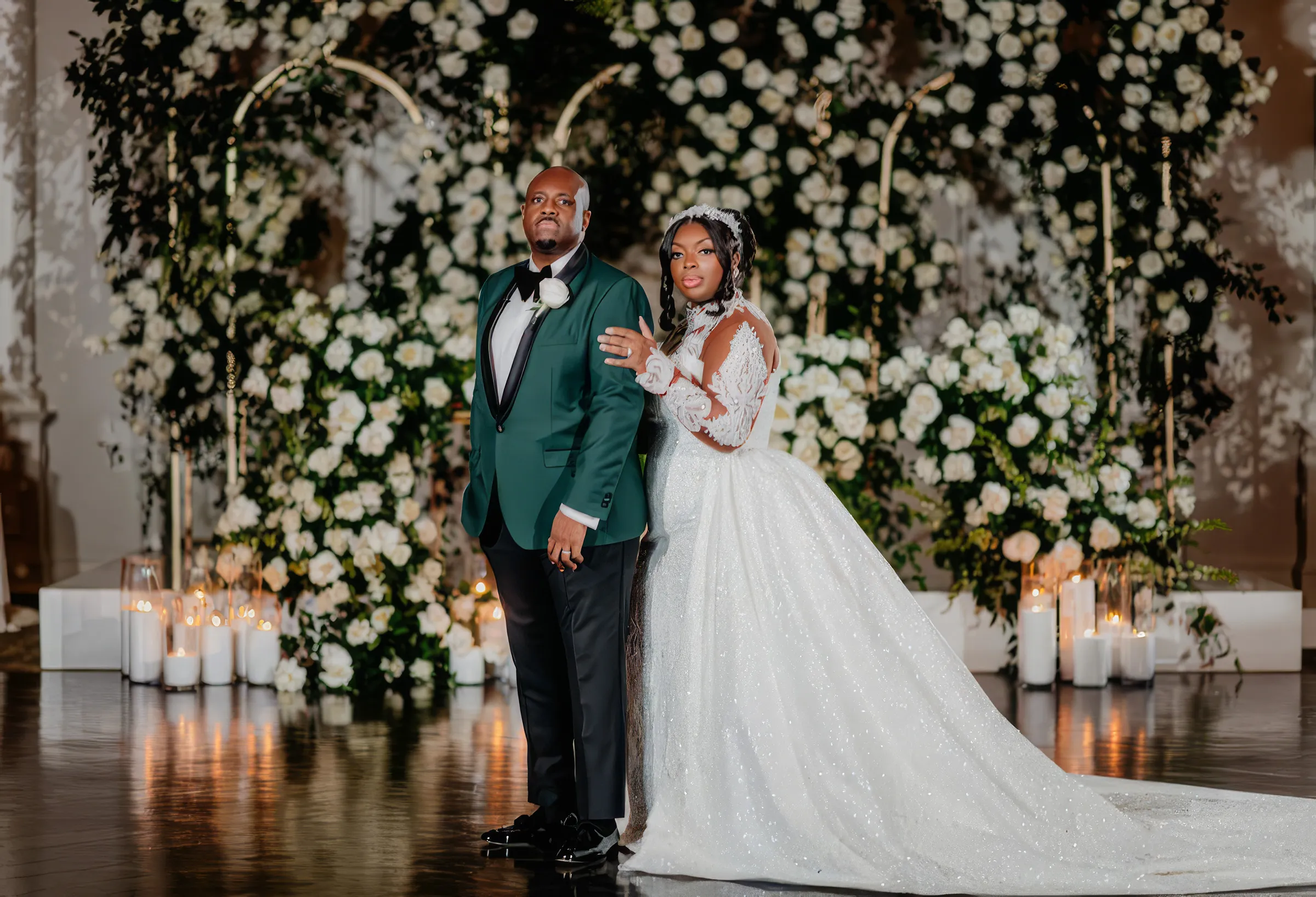 Cathy rests a hand on the chest of her husband, in an emerald tuxedo, among candles and white flowers, her shimmering embellished gown and its long detachable train spread across the reflective floor.