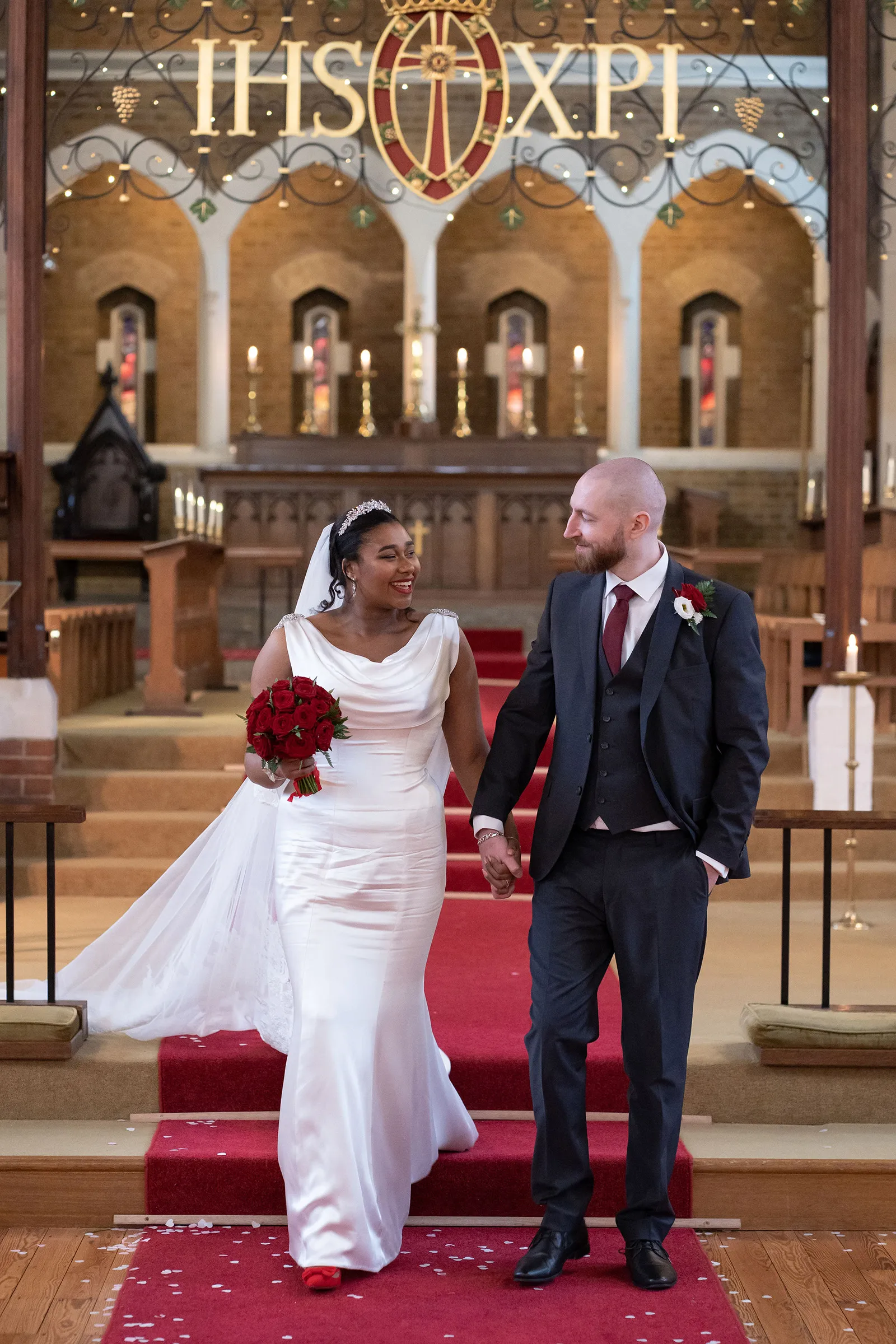 Lola and her husband walk down the red-carpeted steps of a London church. She wears a figure-hugging bespoke Kosibah gown with a cowl neckline and carries red roses, with a lace-edged veil behind her.