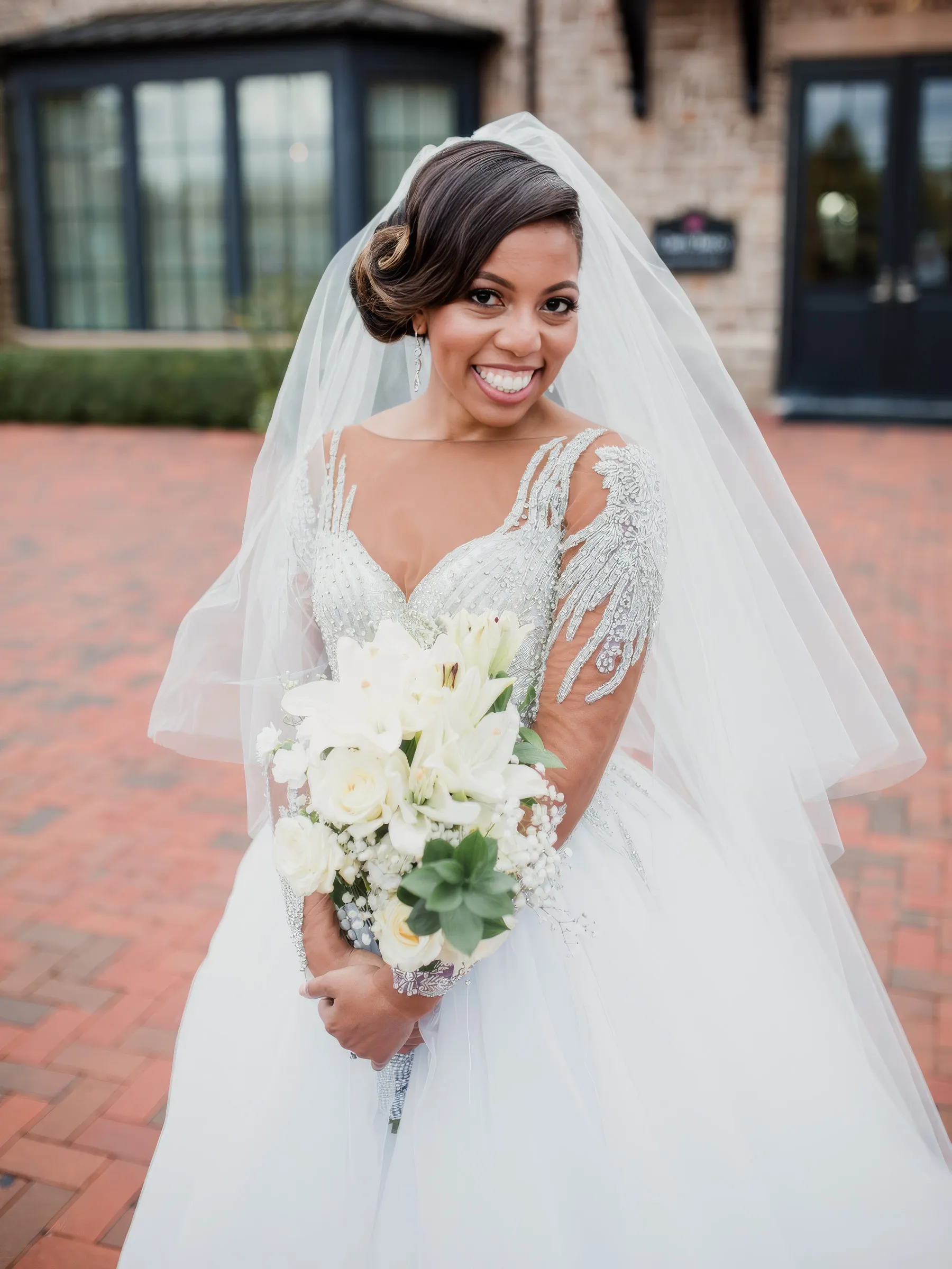 Martina smiles beneath her veil holding white lilies and roses. Her fitted bodice is intricately beaded, and her delicate illusion sleeves are covered in cascading crystal embellishments above a tulle ballgown skirt.