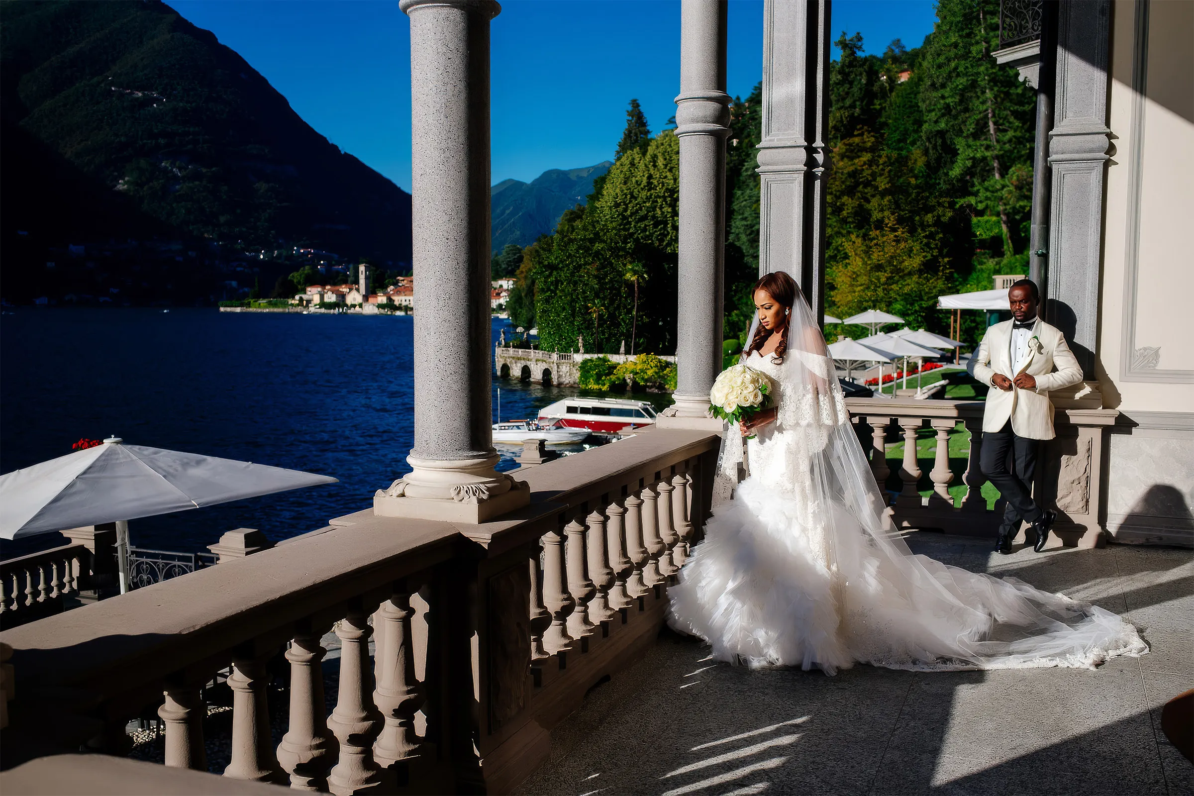 Layal walks along a lakeside terrace at Lake Como, Italy, with her husband in a white dinner jacket leaning on a column behind her. Her ruffled tulle skirt and lace-edged veil trail across the stone.