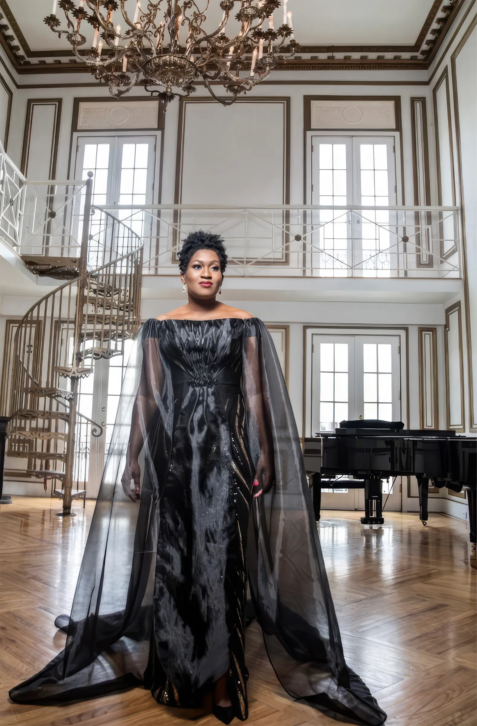 Jackie, New Jersey, standing full length in a sweeping room with a spiral staircase and grand piano, in her black off-the-shoulder Kosibah evening gown, a figure-enhancing silhouette with a floor-length sheer organza cape.