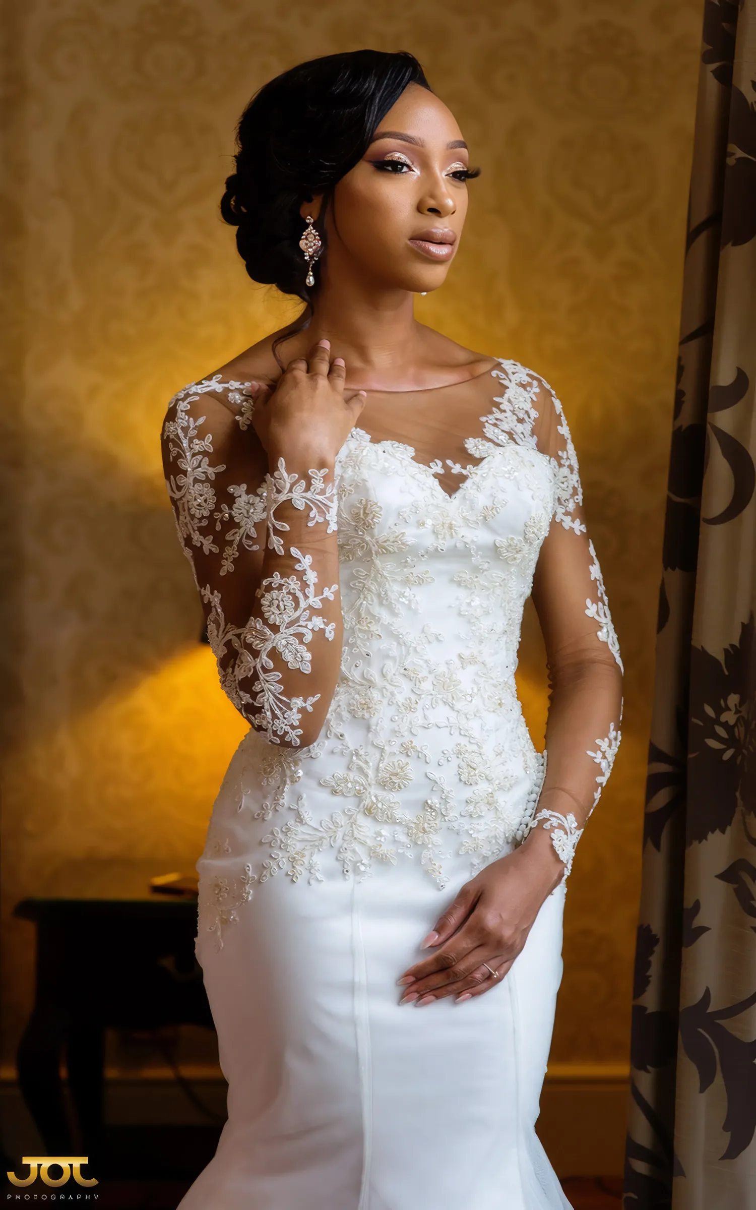 Cherelle poses by a window, one hand at her neck. Delicate floral lace appliqués trail across the illusion neckline, long sheer sleeves and fitted bodice of her figure-hugging Kosibah couture gown.