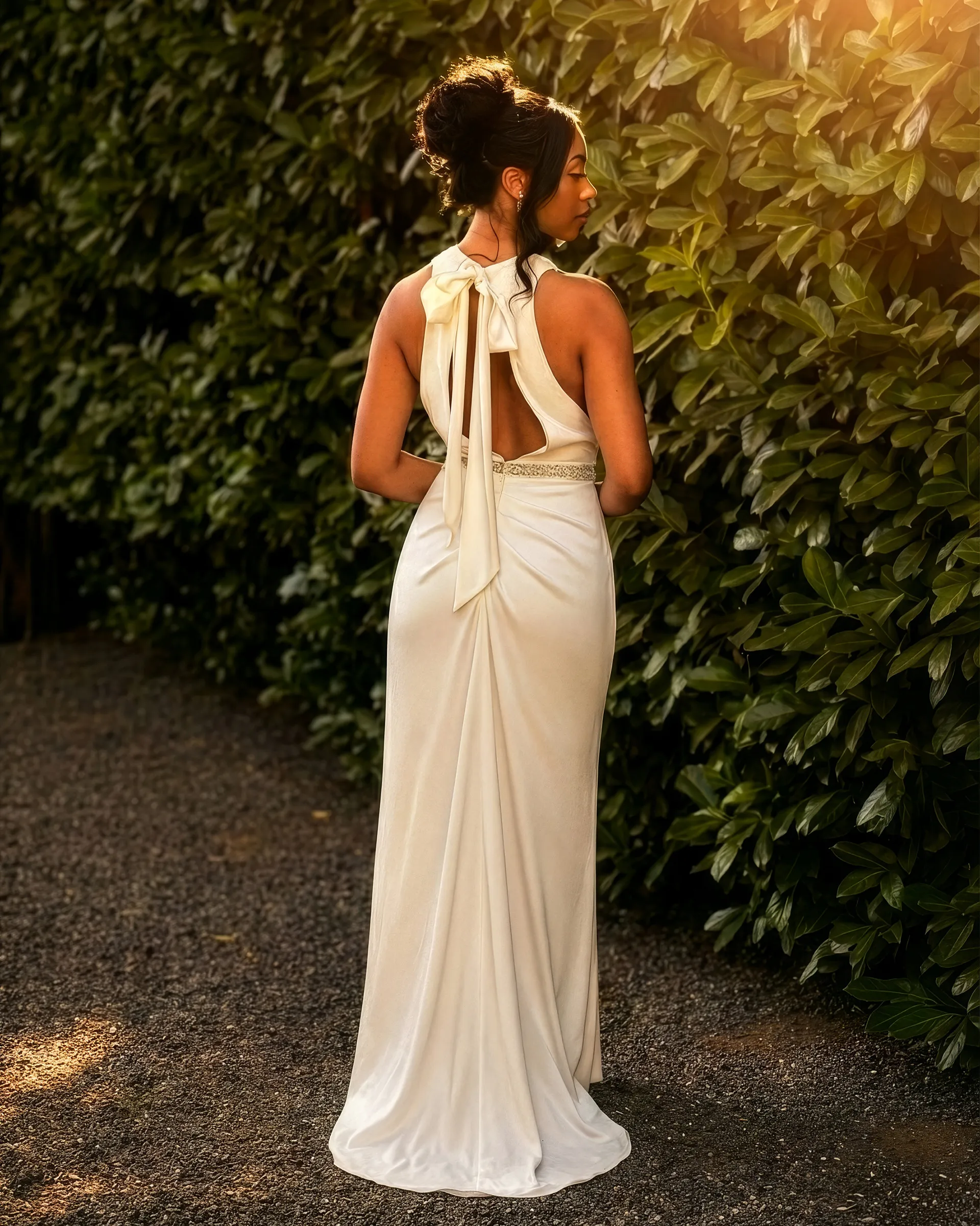 Pearl, back, against a laurel hedge: a satin bow at the nape trails long ribbons over an open keyhole back, above a crystal belt and draping gathered into a short train.
