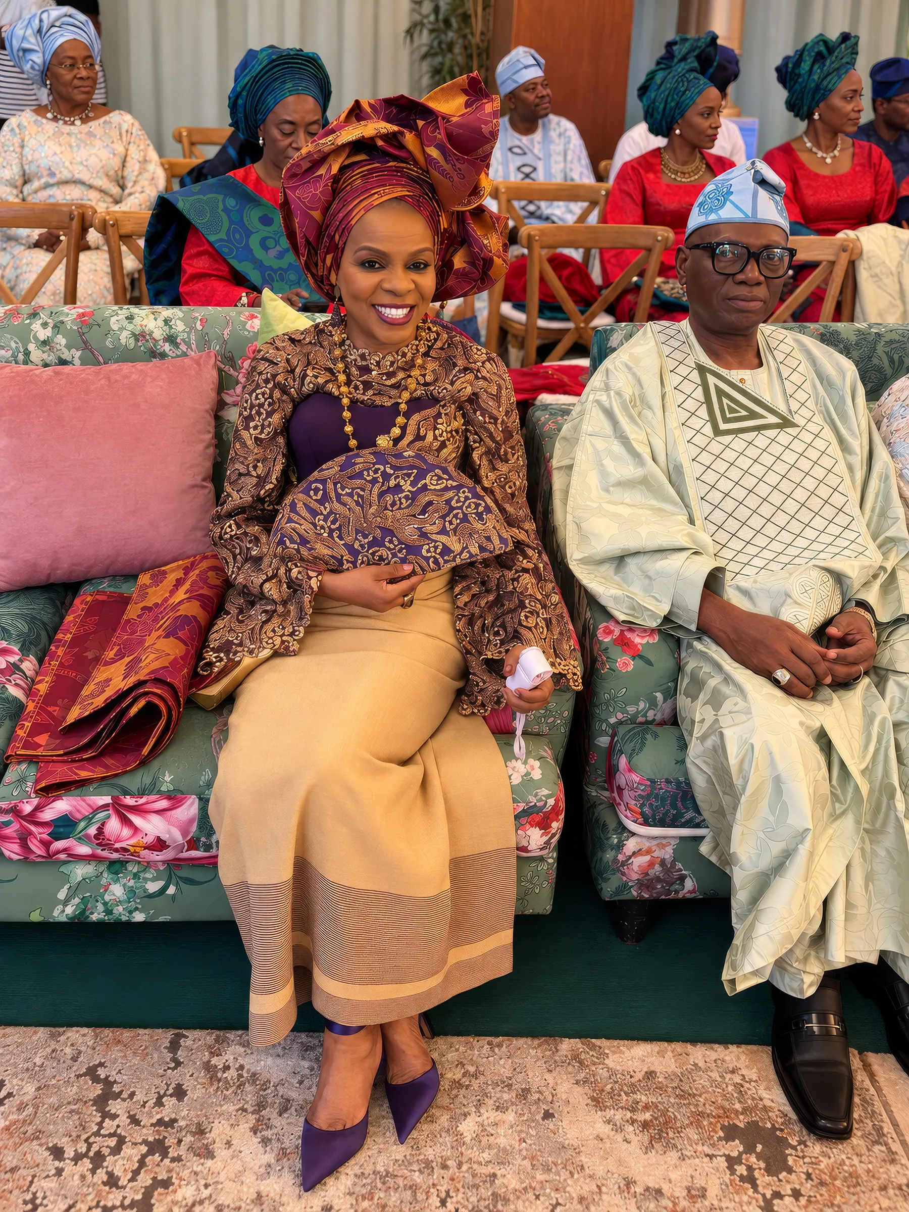 Dupe seated on a floral sofa at the celebration, holding a hand fan, beside a man in a pale-green embroidered agbada and cap; guests in turquoise gele sit behind.