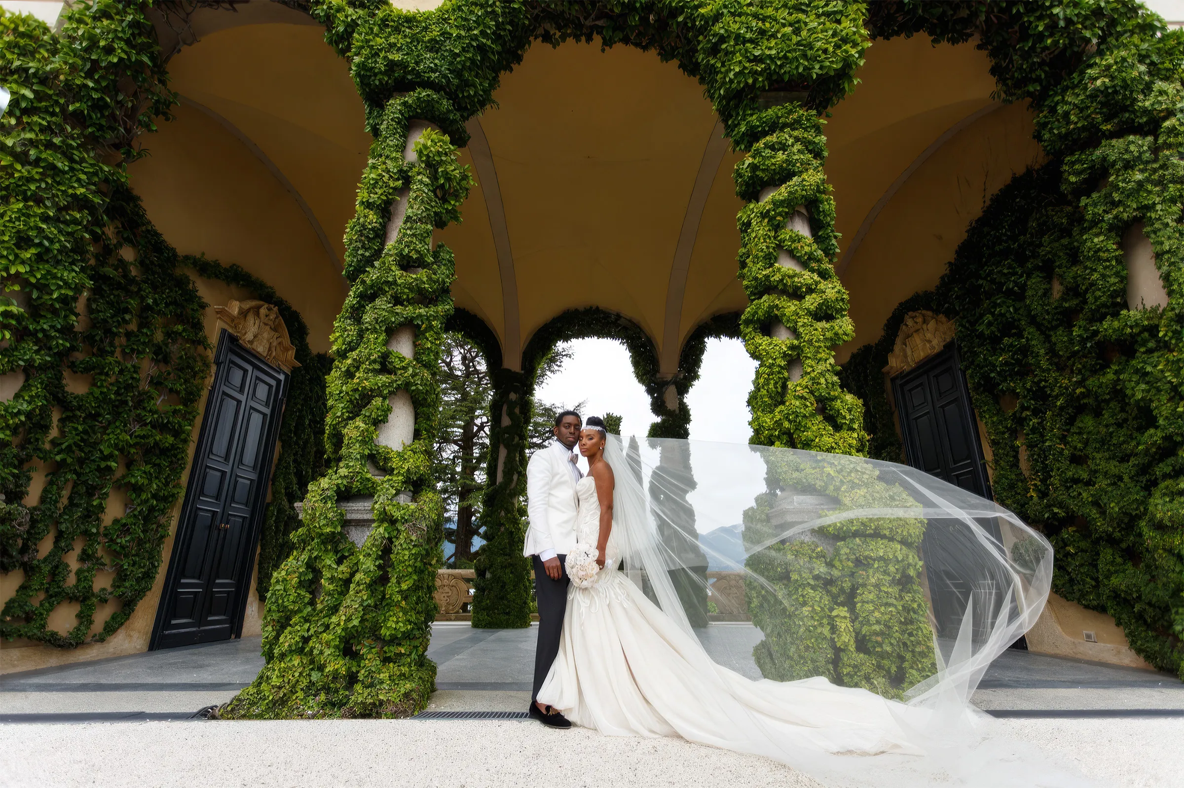 Denise and her husband stand between ivy-covered pillars at Lake Como, Italy, as her cathedral-length veil and the dramatic train of her couture mermaid gown blow out behind her.