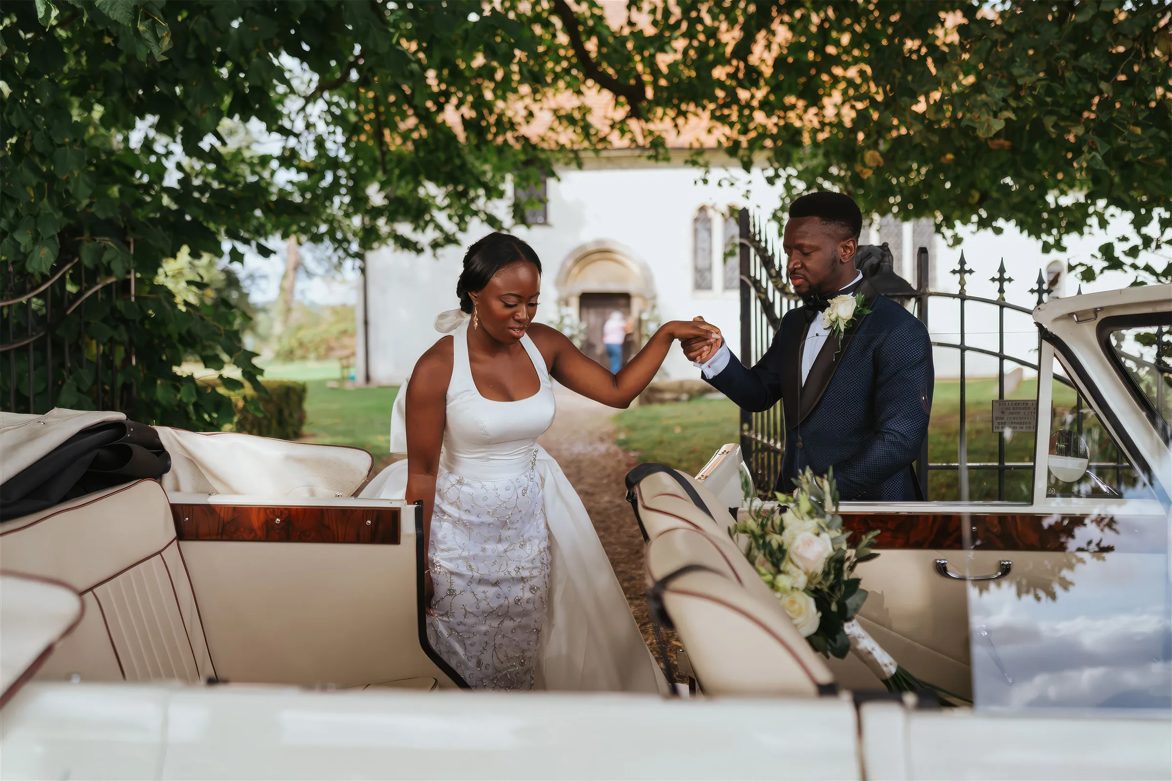 Ife steps out of a vintage cream convertible at Barnston Lodge, Essex, helped by her husband, in a halter-neck bridal gown with a beaded floral-appliquéd skirt and a detachable overskirt.