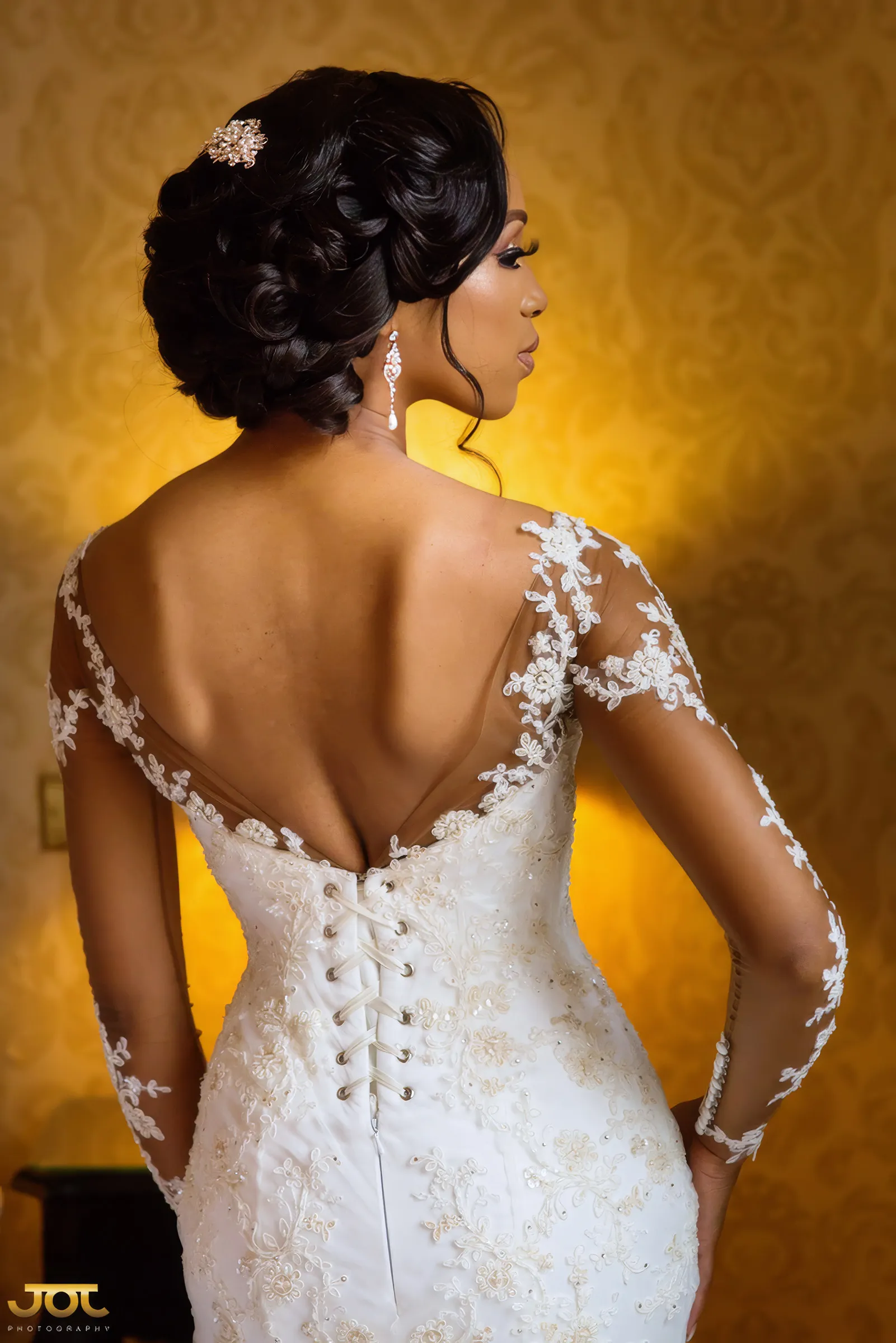 Back view of Cherelle's gown: a low illusion back edged with floral lace appliqués, a row of covered buttons and loops down the bodice, and long sheer lace sleeves. A jewelled pin sits in her swept-up hair.
