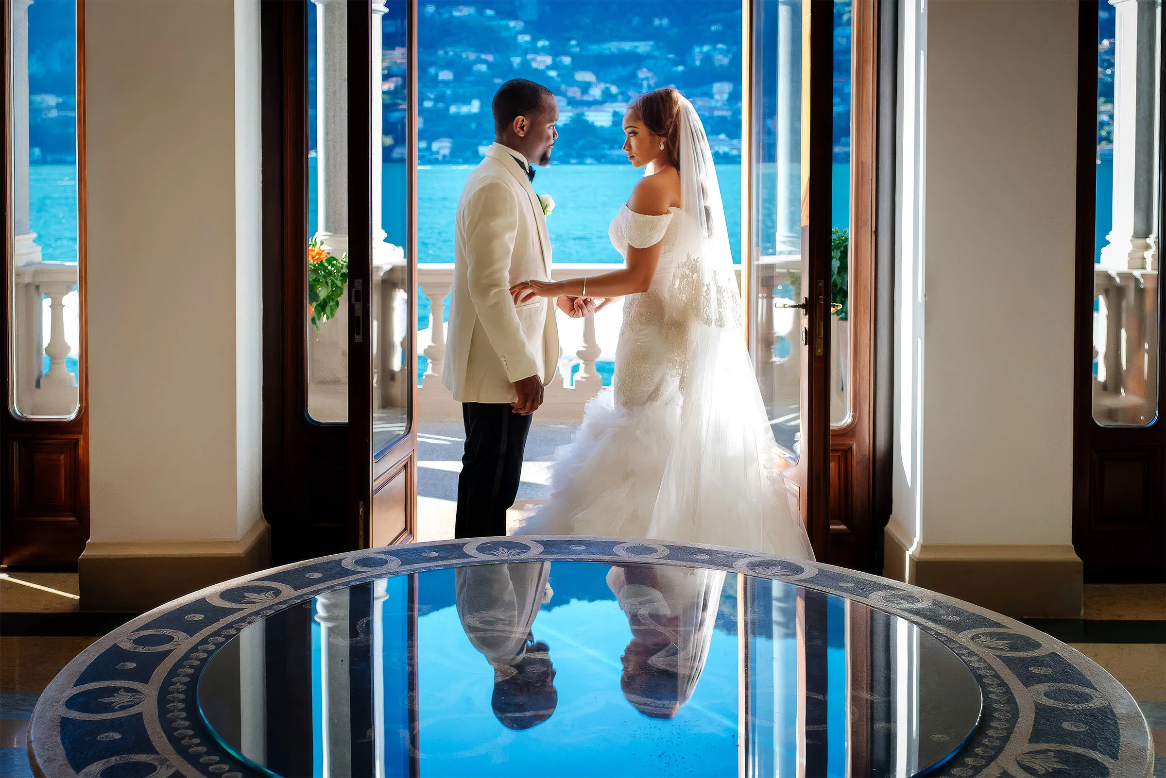 Layal and her husband face each other on a terrace above Lake Como, Italy, their reflections in a glass table. Her off-the-shoulder gown has a sculpted bodice and a ruffled tulle skirt.