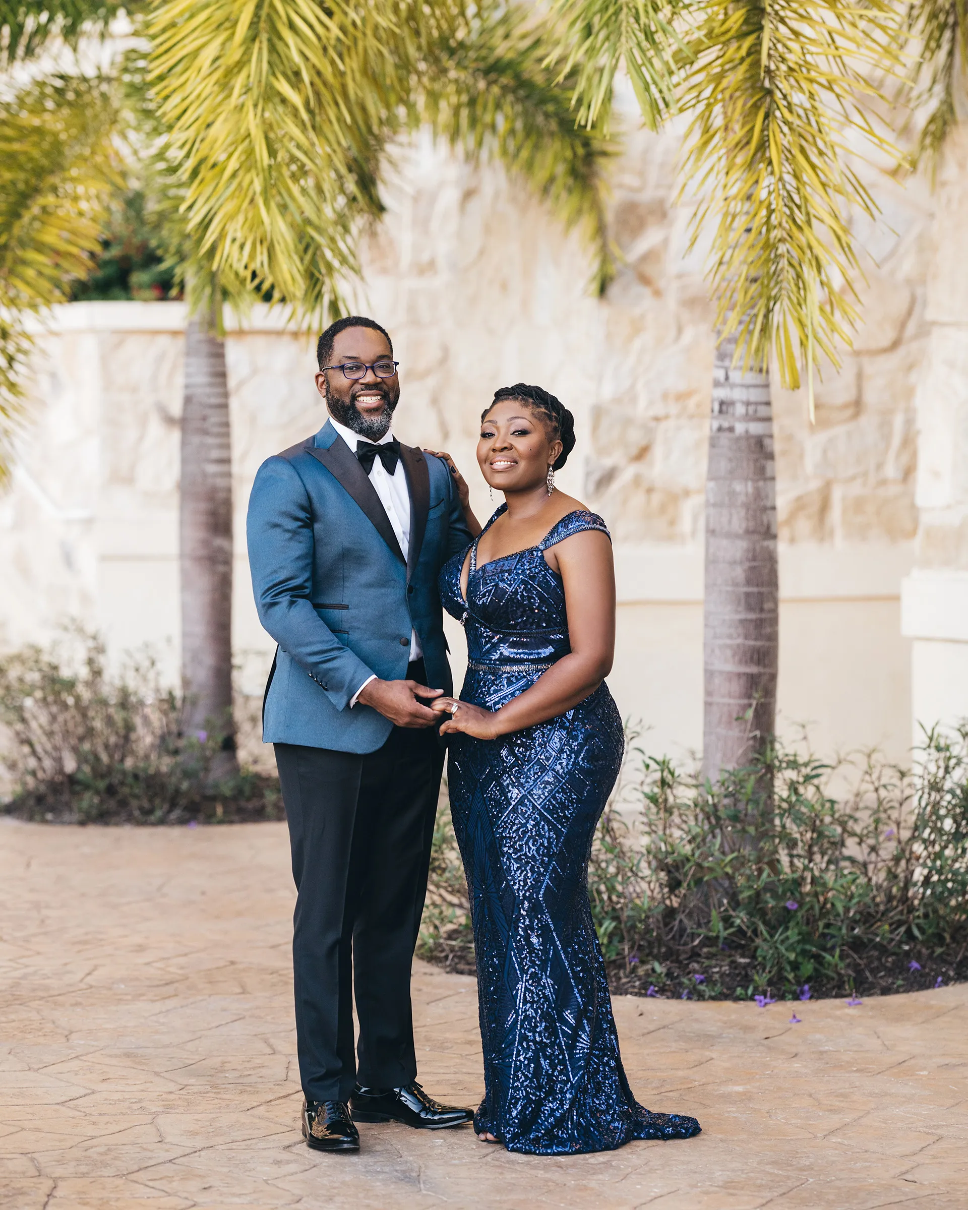 Nana and her husband pose in front of palm trees. Nana wears a floor-length Kosibah evening wear gown in midnight-blue sequinned lace with beaded straps and a subtle train, shaping an hourglass silhouette.