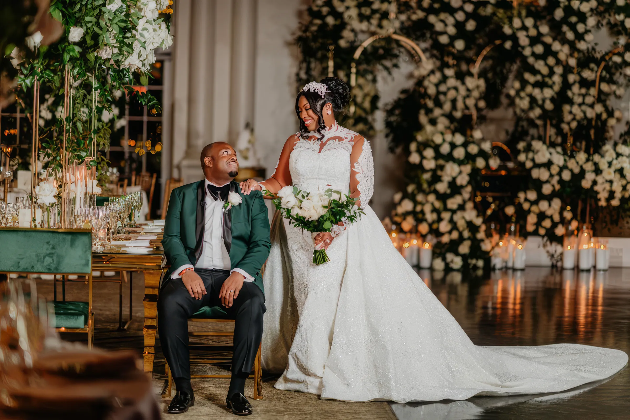 Cathy stands holding white roses beside her husband, seated in an emerald tuxedo. The embroidered floral motifs and lace sleeves of her fitted gown catch the candlelight, its detachable train spread behind her.