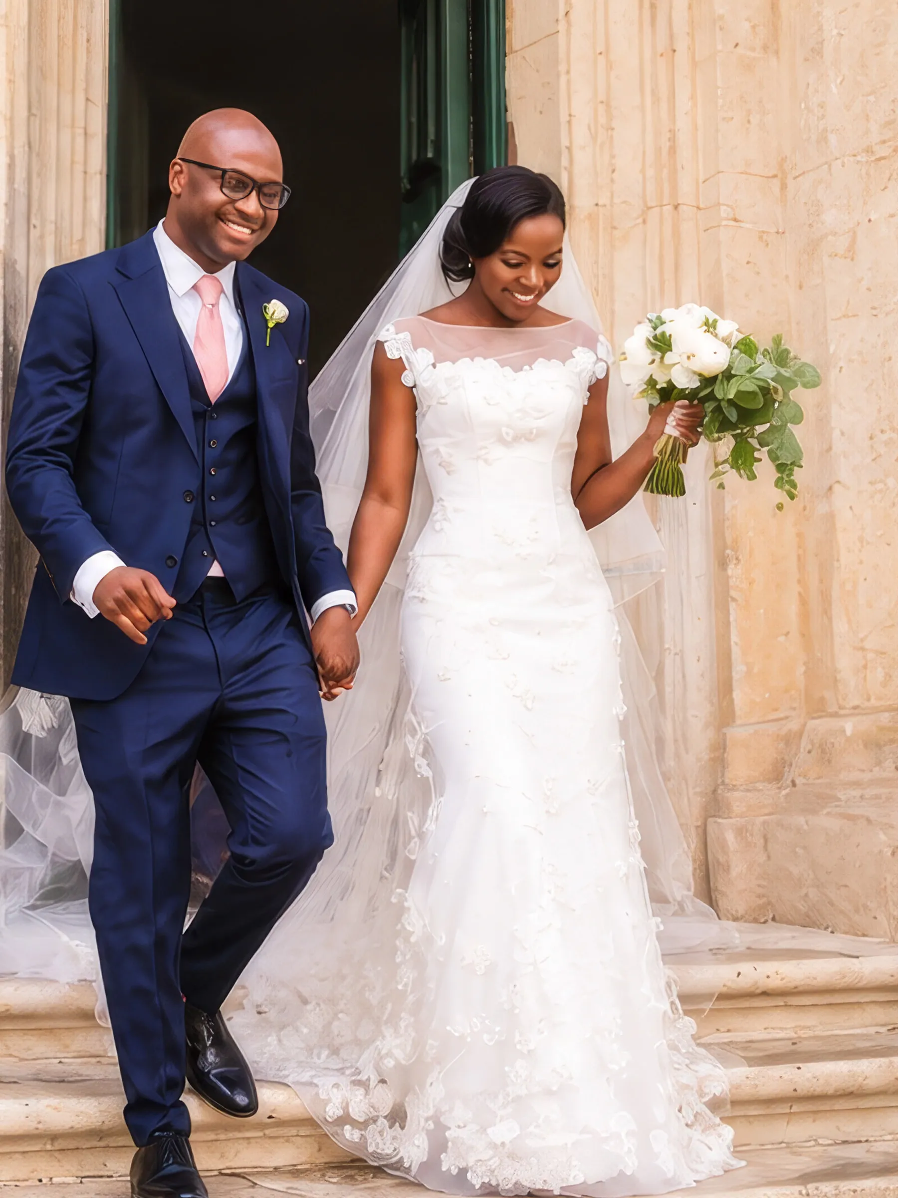 Clara and her husband walk hand in hand down the stone steps after their ceremony in Valletta, Malta. Her fitted gown has an illusion neckline and floral appliqués, with a long tulle veil and a bouquet of white roses.
