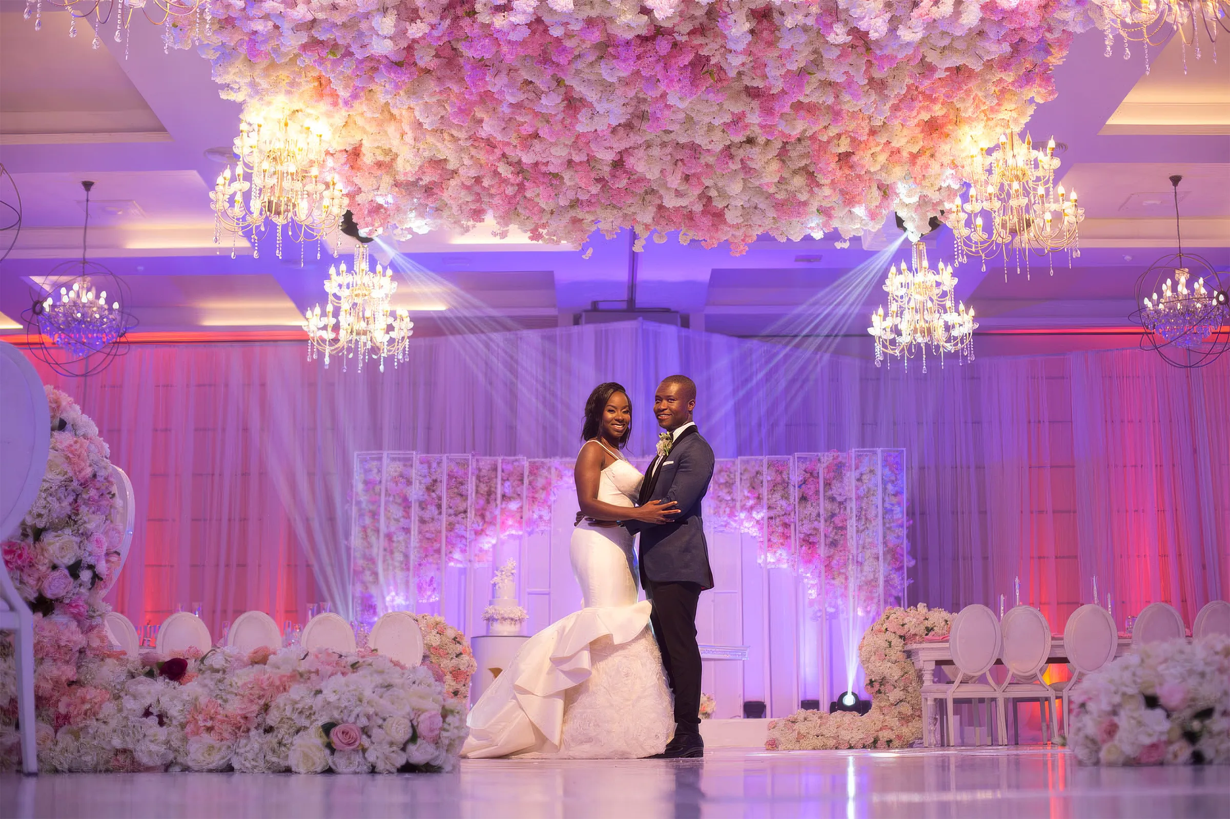 Nana Ama and her husband share their first dance beneath a ceiling of pink and white flowers and crystal chandeliers. Her fitted gown blossoms into a dramatic trumpet skirt with architectural folds and three-dimensional floral textures.