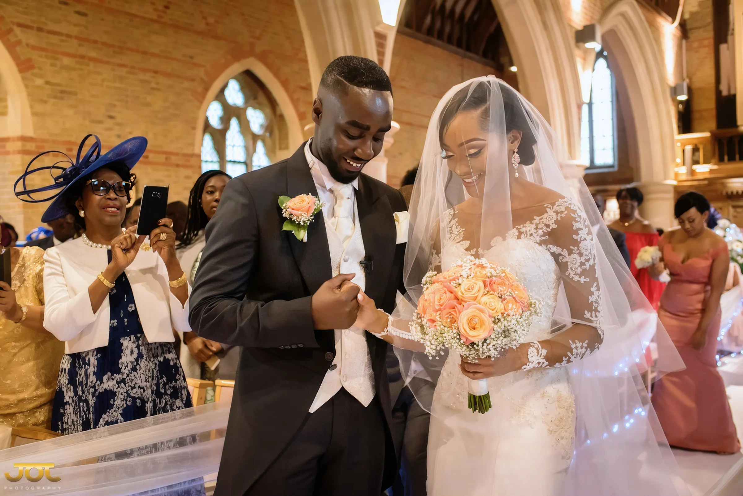 Cherelle smiles at her husband as they exchange rings during their church ceremony in London. Her gown has an illusion neckline and long sheer sleeves covered in floral lace appliqués, with a long tulle veil and peach roses.