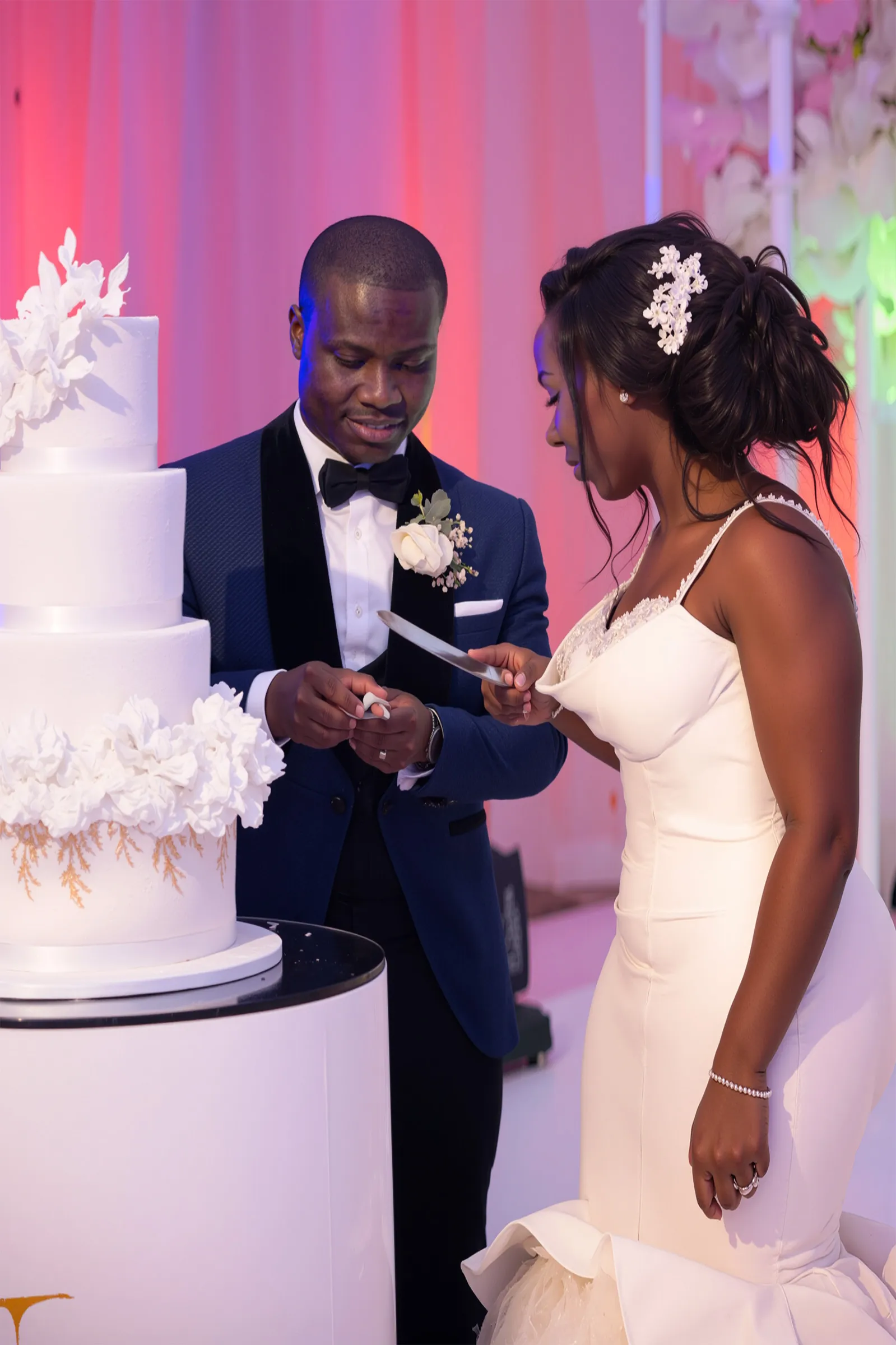 Nana Ama and her husband cut their tiered white wedding cake, decorated with sugar flowers. Her figure-hugging gown has a draped cowl neckline and delicately embellished shoulder straps, with a jewelled ornament in her hair.