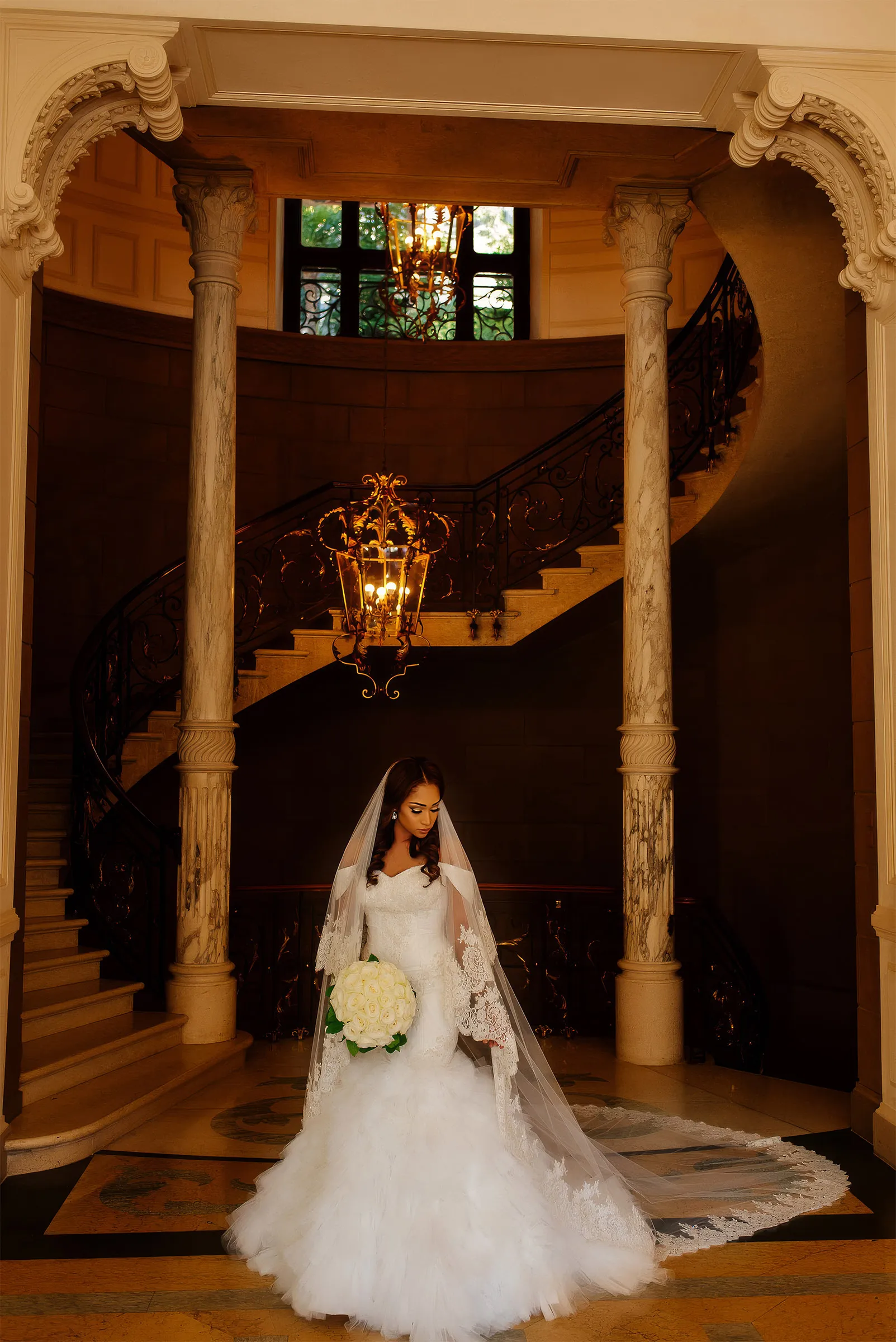 Layal, in her couture gown with a ruffled tulle skirt and a long lace-edged veil, looks down at her bouquet of cream roses beneath a chandelier at the foot of a grand staircase.