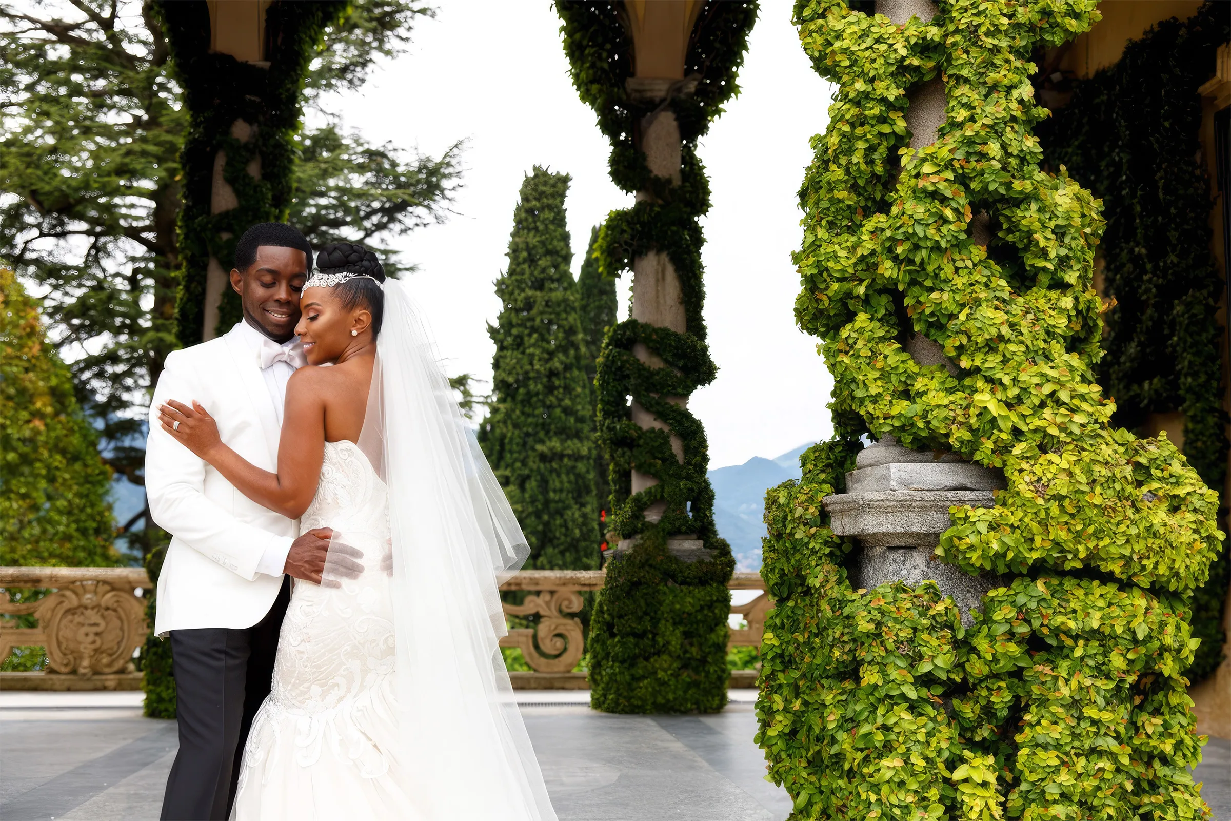 Denise and her husband embrace on a terrace at Lake Como, Italy. Her strapless mermaid gown is fitted through the bodice, with a long sheer veil falling down her back.
