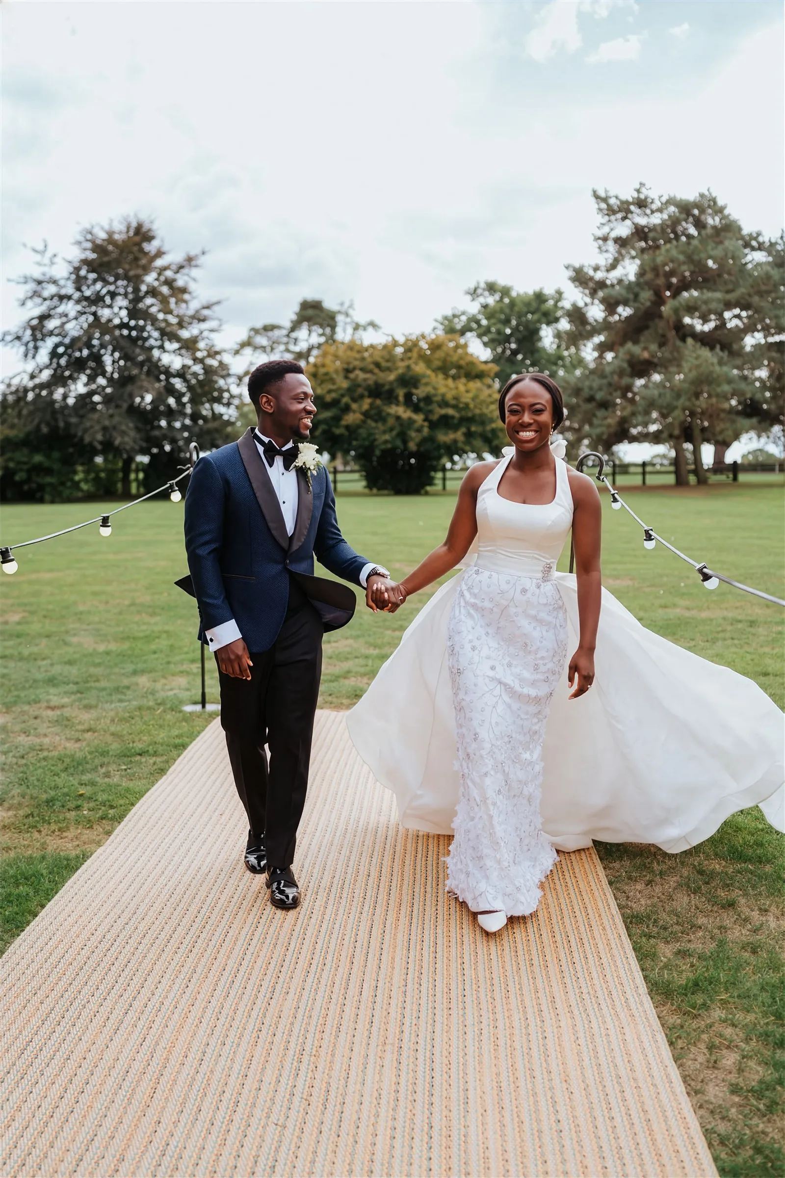 Ife and her husband walk hand in hand across the lawn at Barnston Lodge, Essex, laughing. Her halter-neck gown has an embellished fitted skirt, with the detachable overskirt billowing out behind her.
