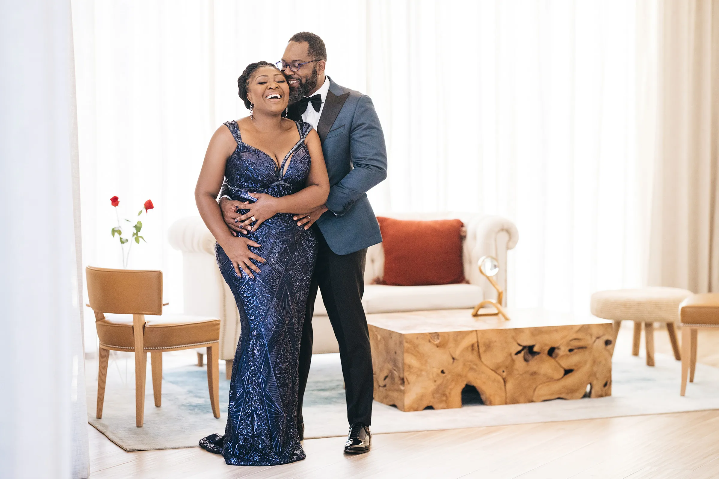 Nana laughs as her husband embraces her from behind in a bright sitting room. Her midnight-blue sequinned lace Kosibah gown has a plunging neckline and a defined, contoured waist.