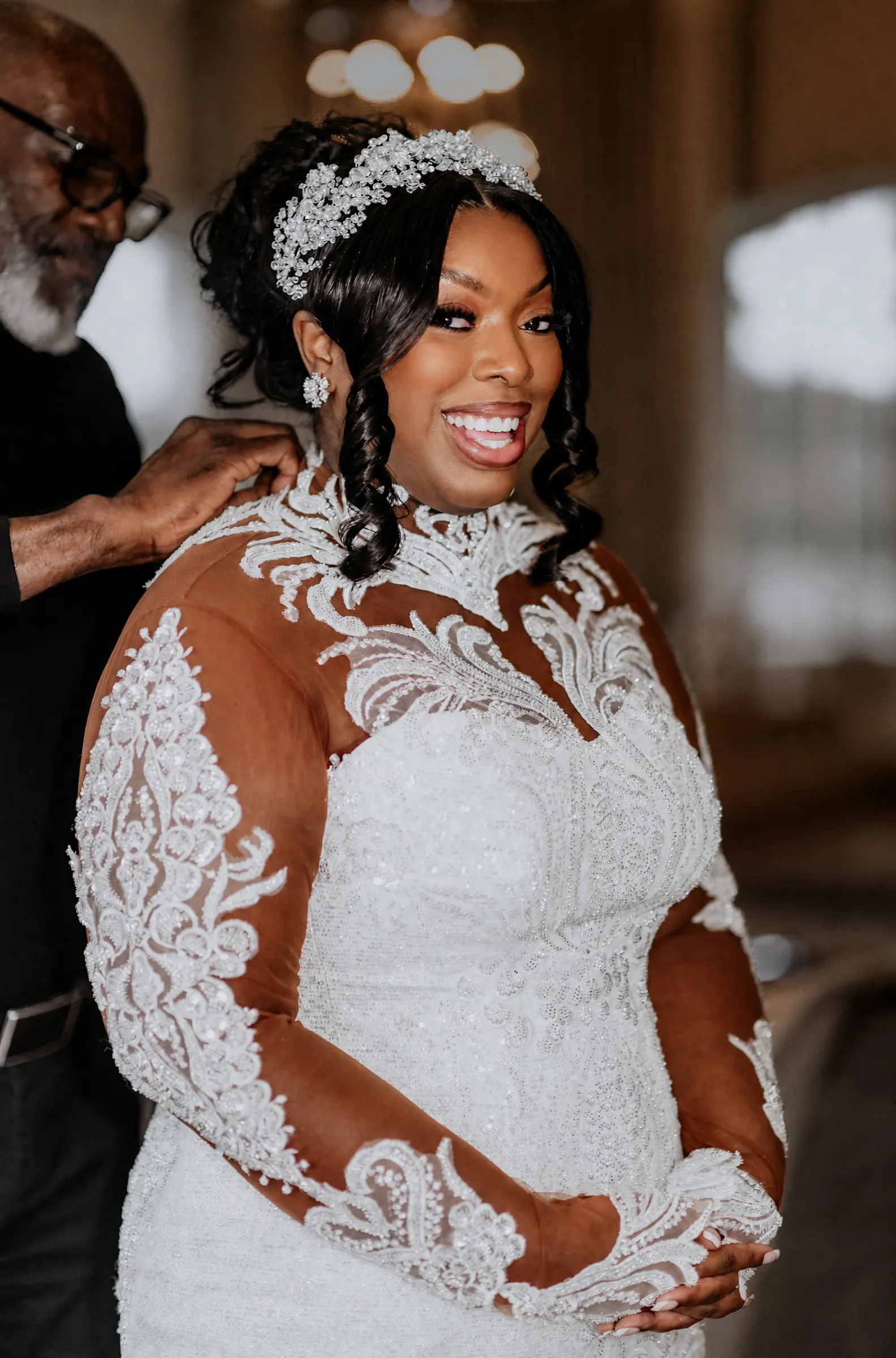 Cathy smiles beneath a jewelled tiara as Yemi fastens the back of her gown. Close up, lace appliqués climb the high illusion neckline and long sheer sleeves over a shimmering embellished bodice.