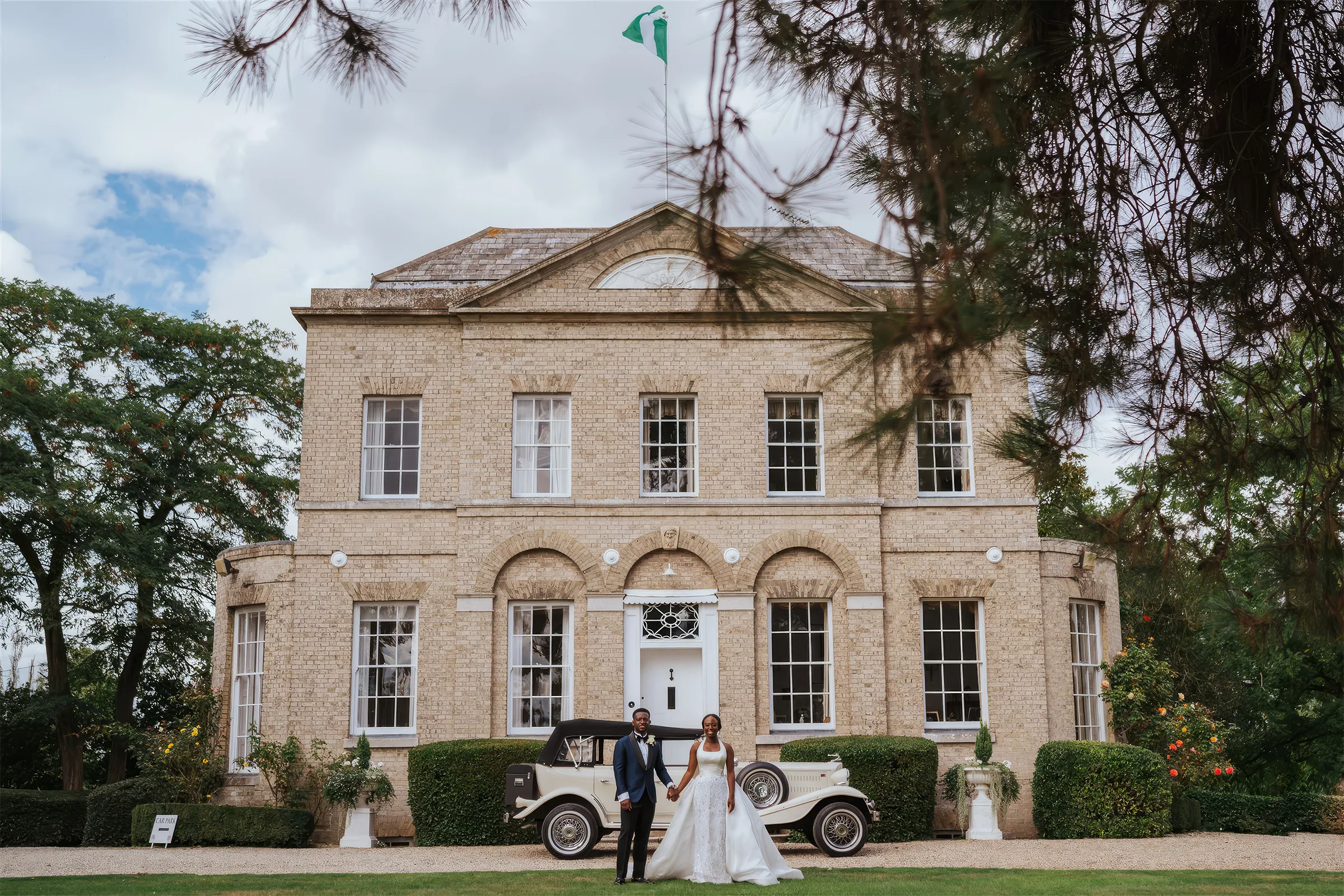 Ife and her husband stand in front of the Georgian manor house at Barnston Lodge, Essex, beside a vintage open-top car. She wears a bespoke Kosibah couture bridal gown with a flowing train.