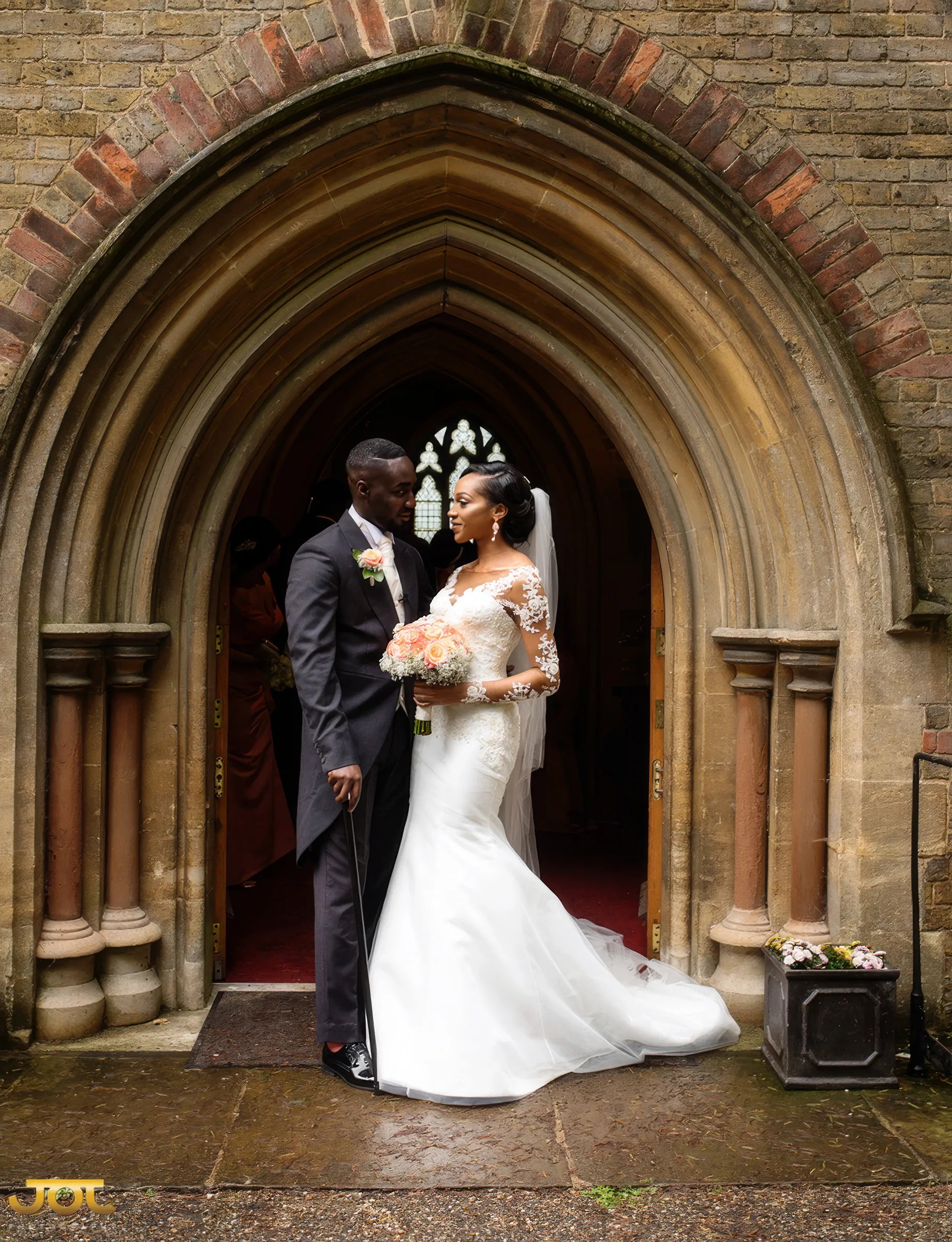 Cherelle and her husband stand in an arched stone church doorway. Her sculpted, figure-hugging gown flows into a flared skirt and train, with lace appliqués on the illusion neckline and long sheer sleeves.