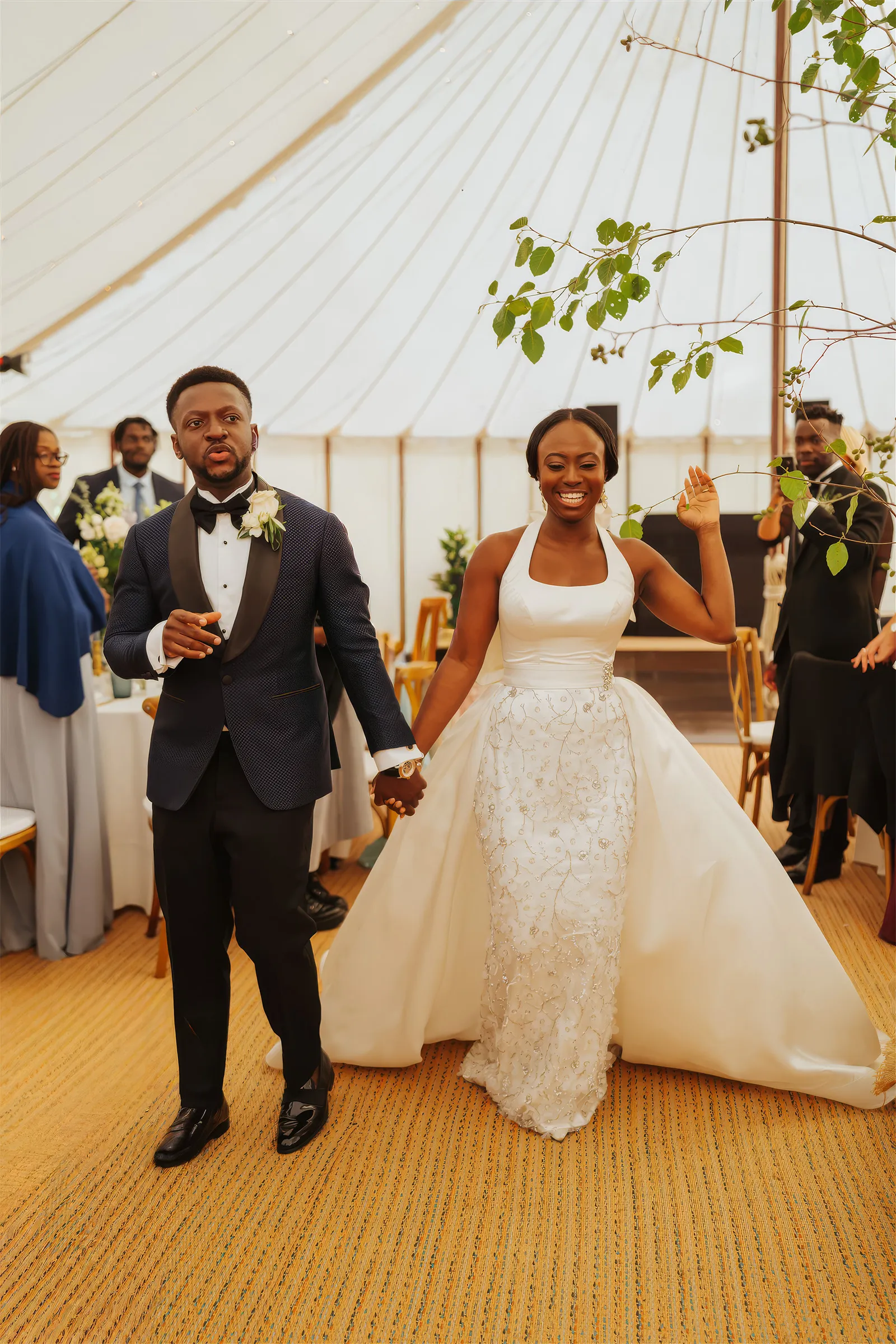 Ife and her husband enter the marquee reception at Barnston Lodge, Essex, hand in hand. She raises an arm in a halter-neck gown with an embellished floral skirt and detachable overskirt.
