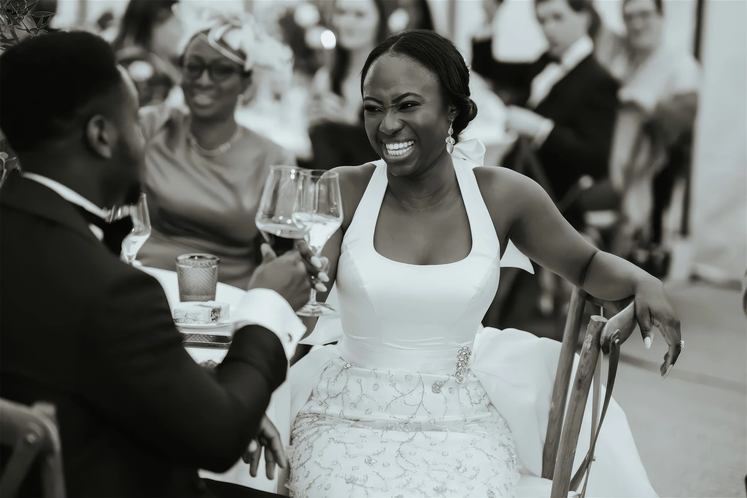 Black and white close-up of Ife laughing as she toasts with her husband. Her white halter-neck bodice is edged with a beaded sash above a delicately embellished skirt.