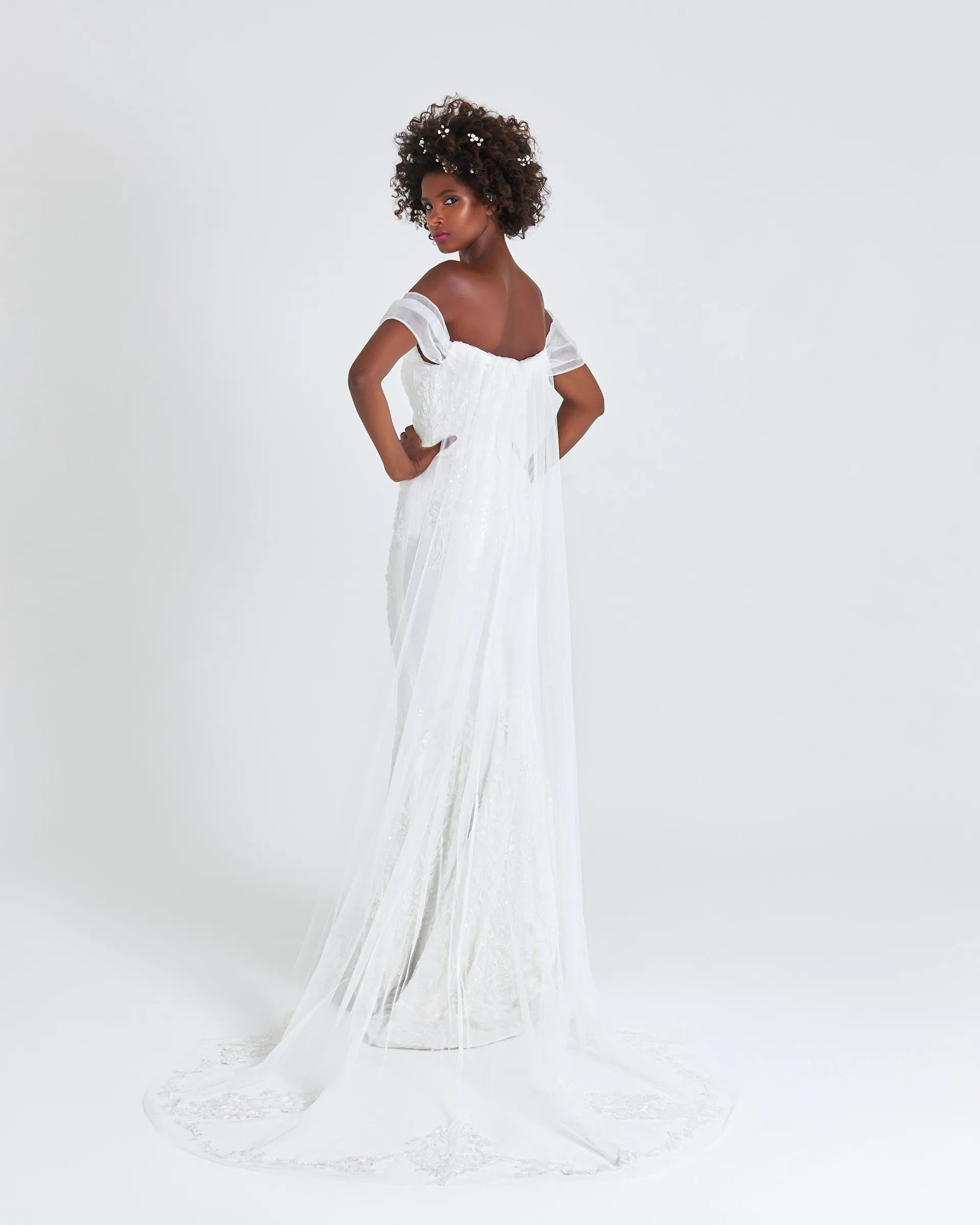 Ajoke, back, looking over her shoulder: the Watteau train flows from the off-the-shoulder sleeves to a cathedral-length hem edged with lace appliqués.