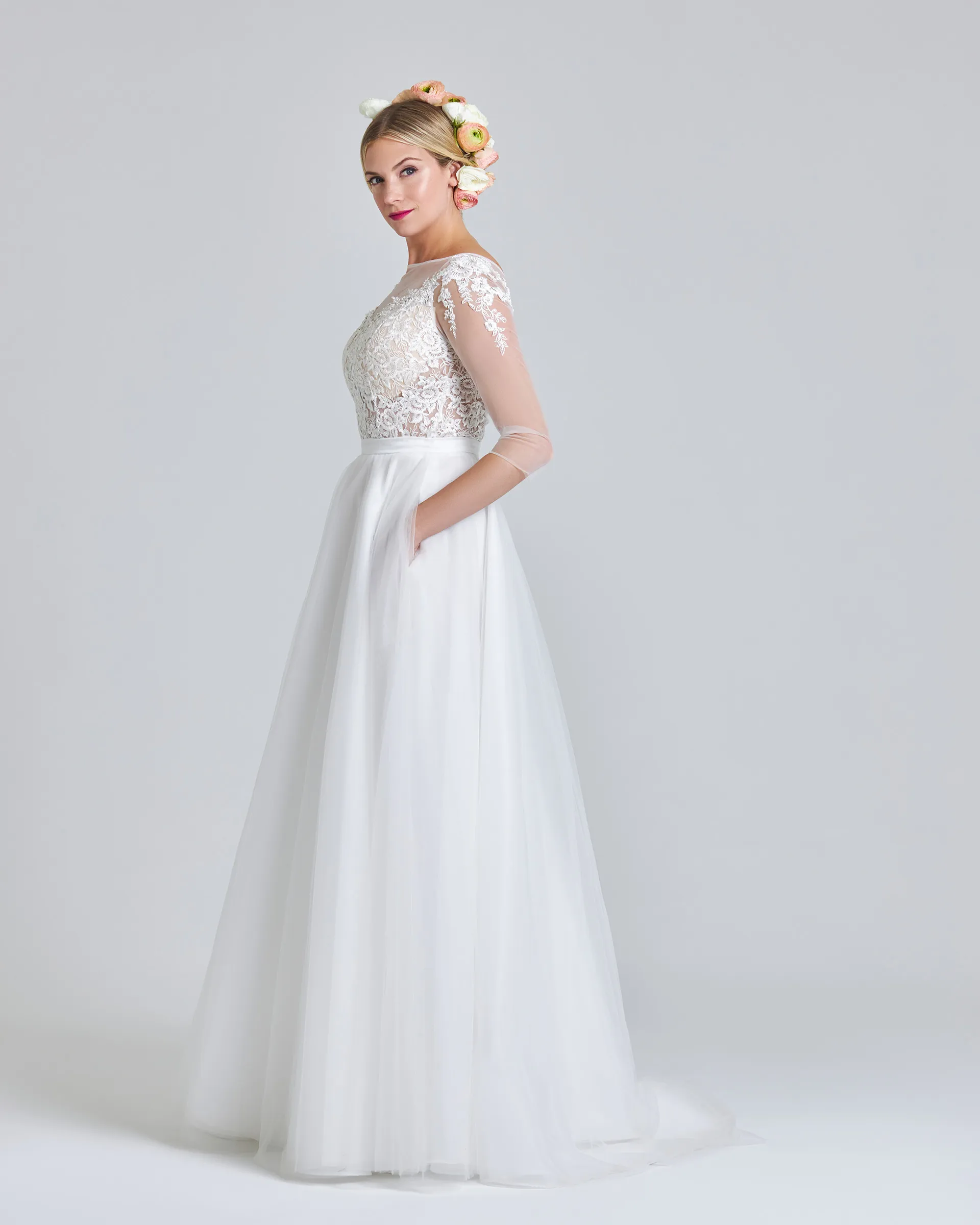 Ariyike, side, hand in a pocket of the full tulle skirt, which falls to a short train; lace appliqués trail down the sheer sleeve.