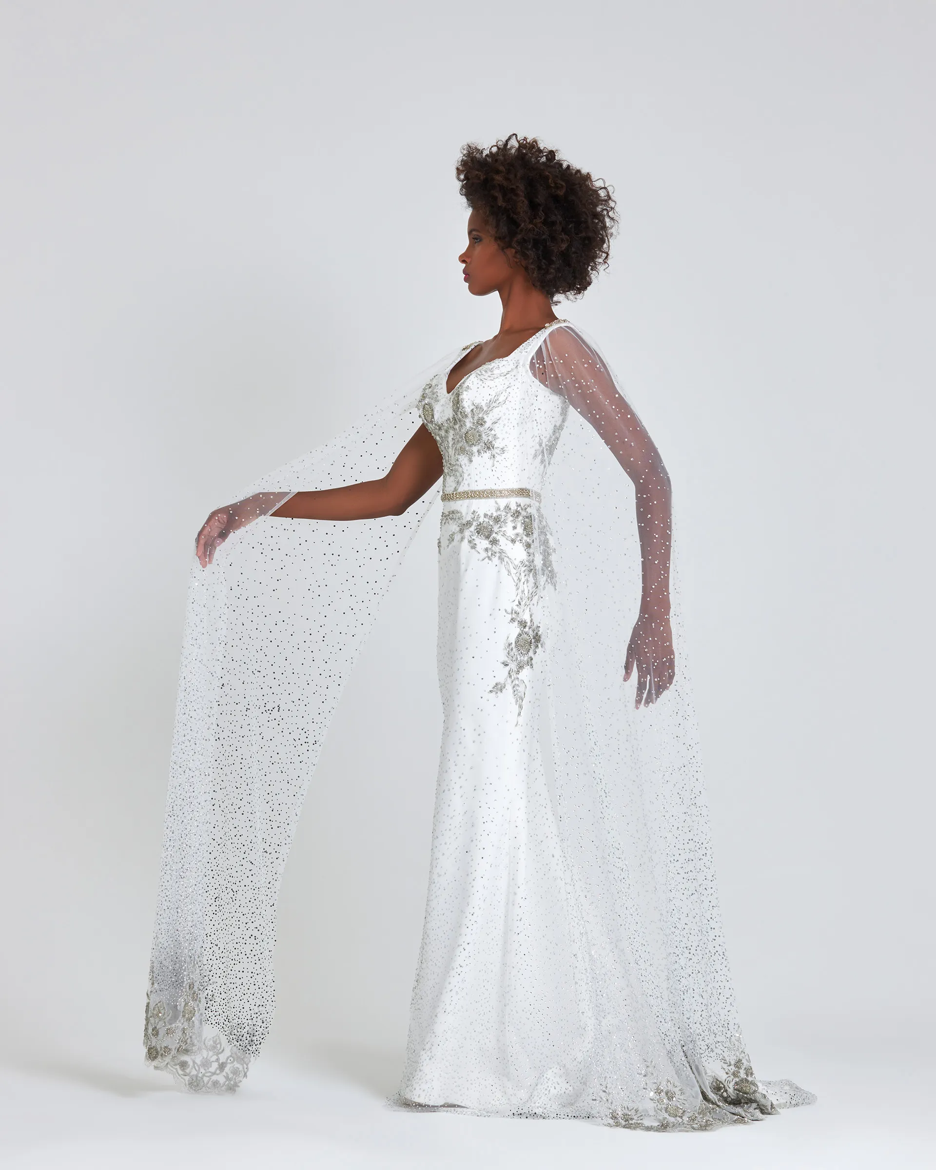 Ashake, side, one arm lifted to spread a cape sleeve: crystal-studded tulle drifting to the floor, embroidered at its hem.