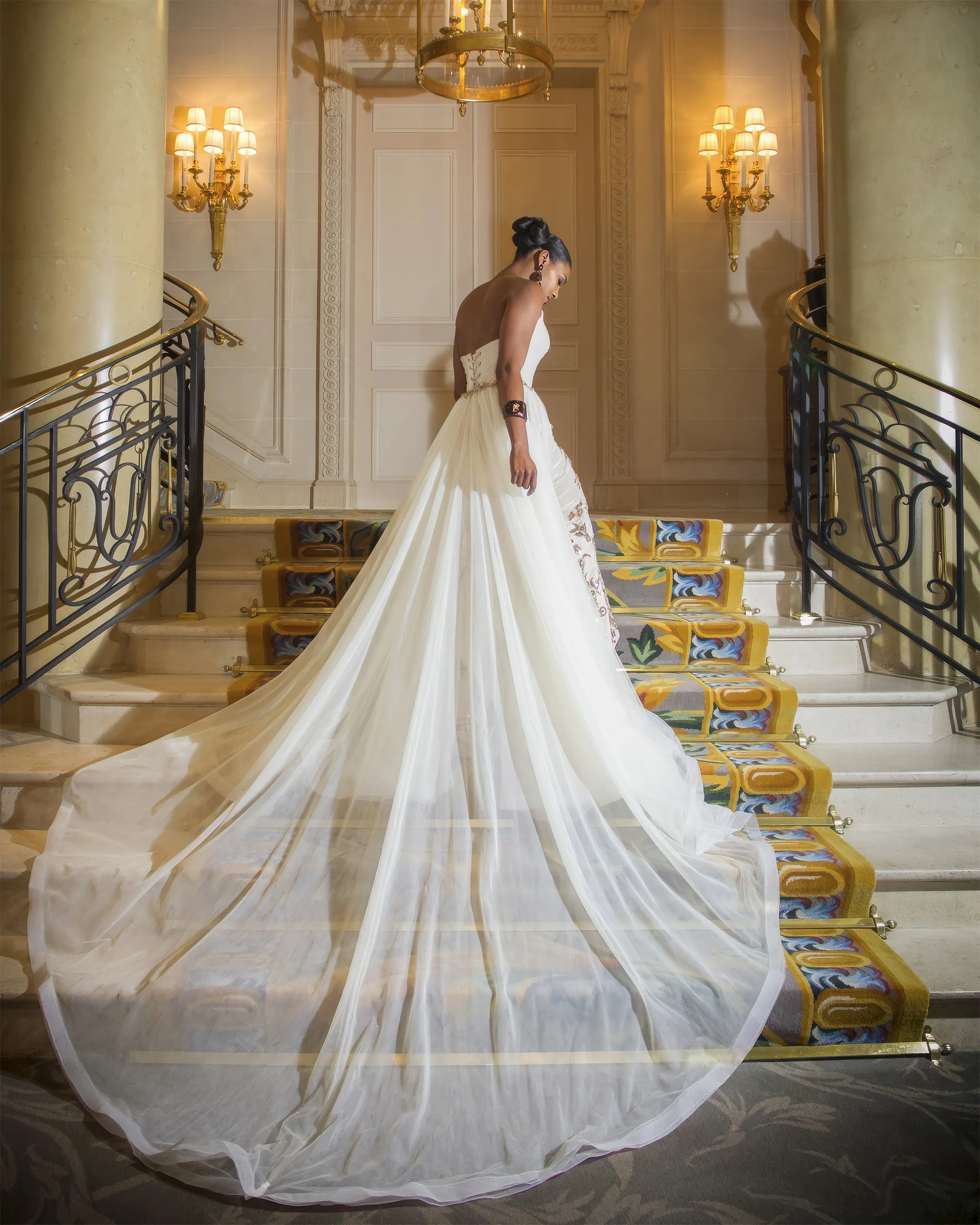 Calliope from behind at the foot of a marble staircase, the tulle overskirt fanning out into a cathedral train across the steps.