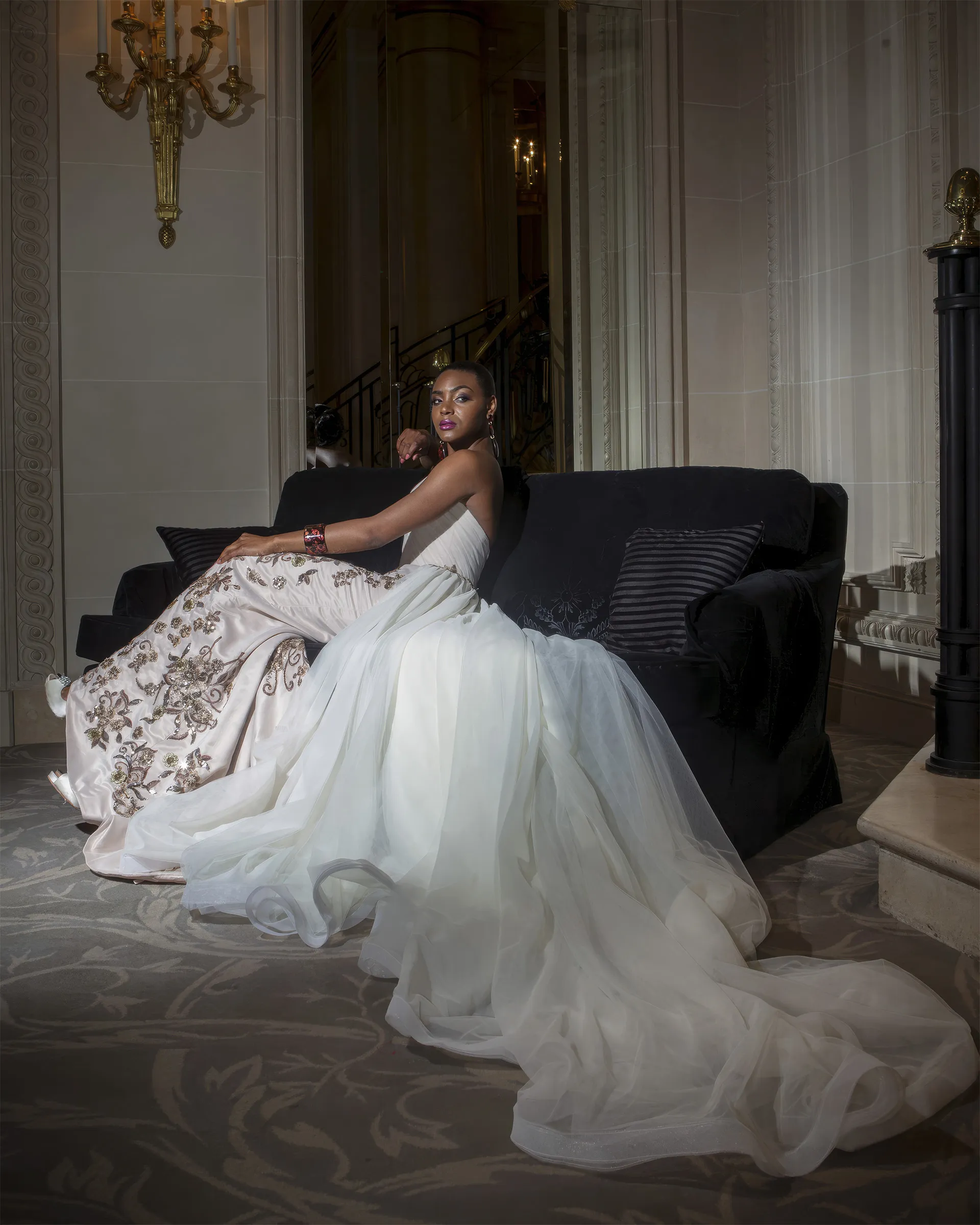 Calliope seated on a black velvet sofa in a grand hotel lobby, the bronze appliqués on the column skirt glinting and the tulle train spread around her.
