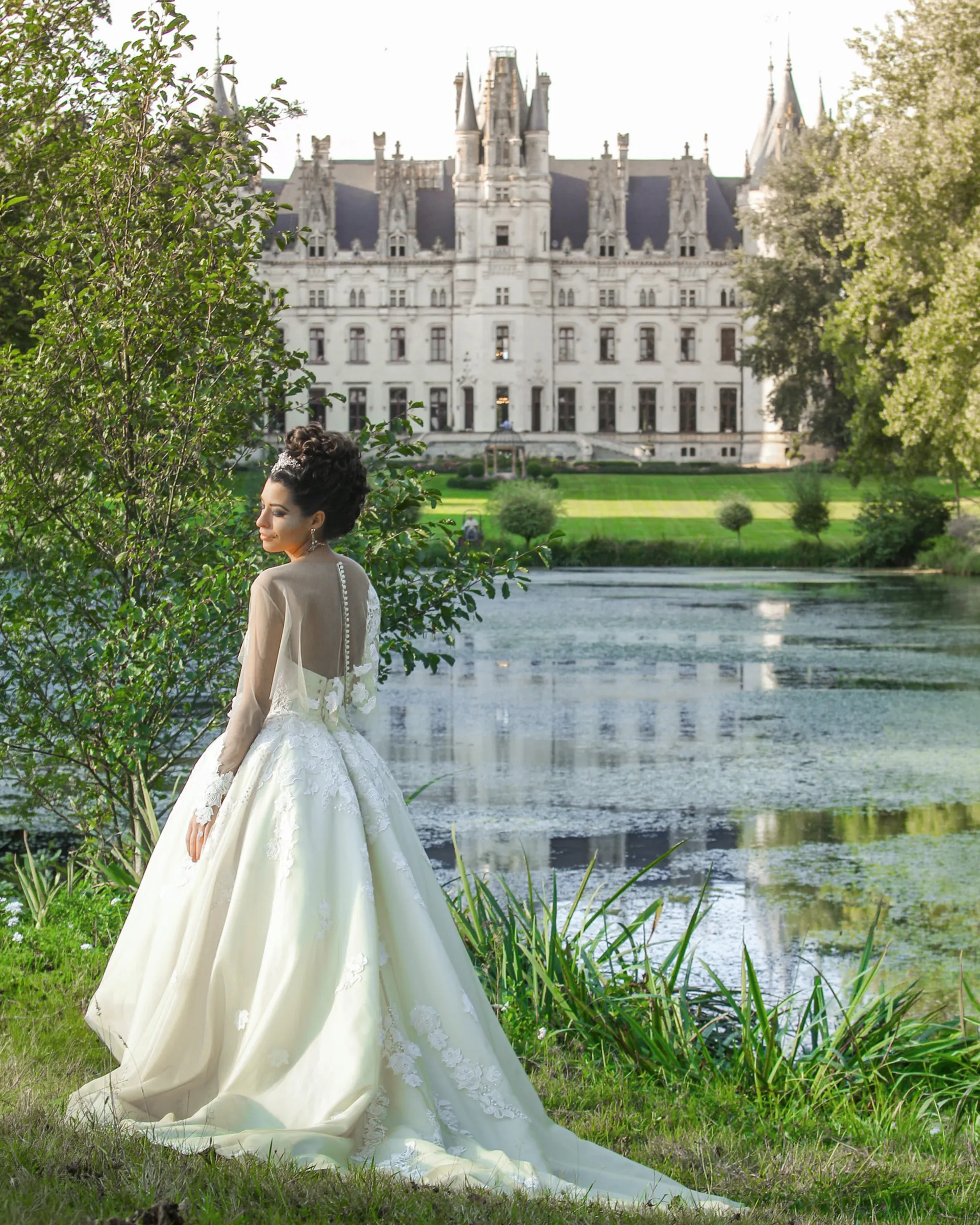 Clio from behind, facing the château across its lake: covered buttons down the sheer jacket and the layered tulle skirt falling into a train.