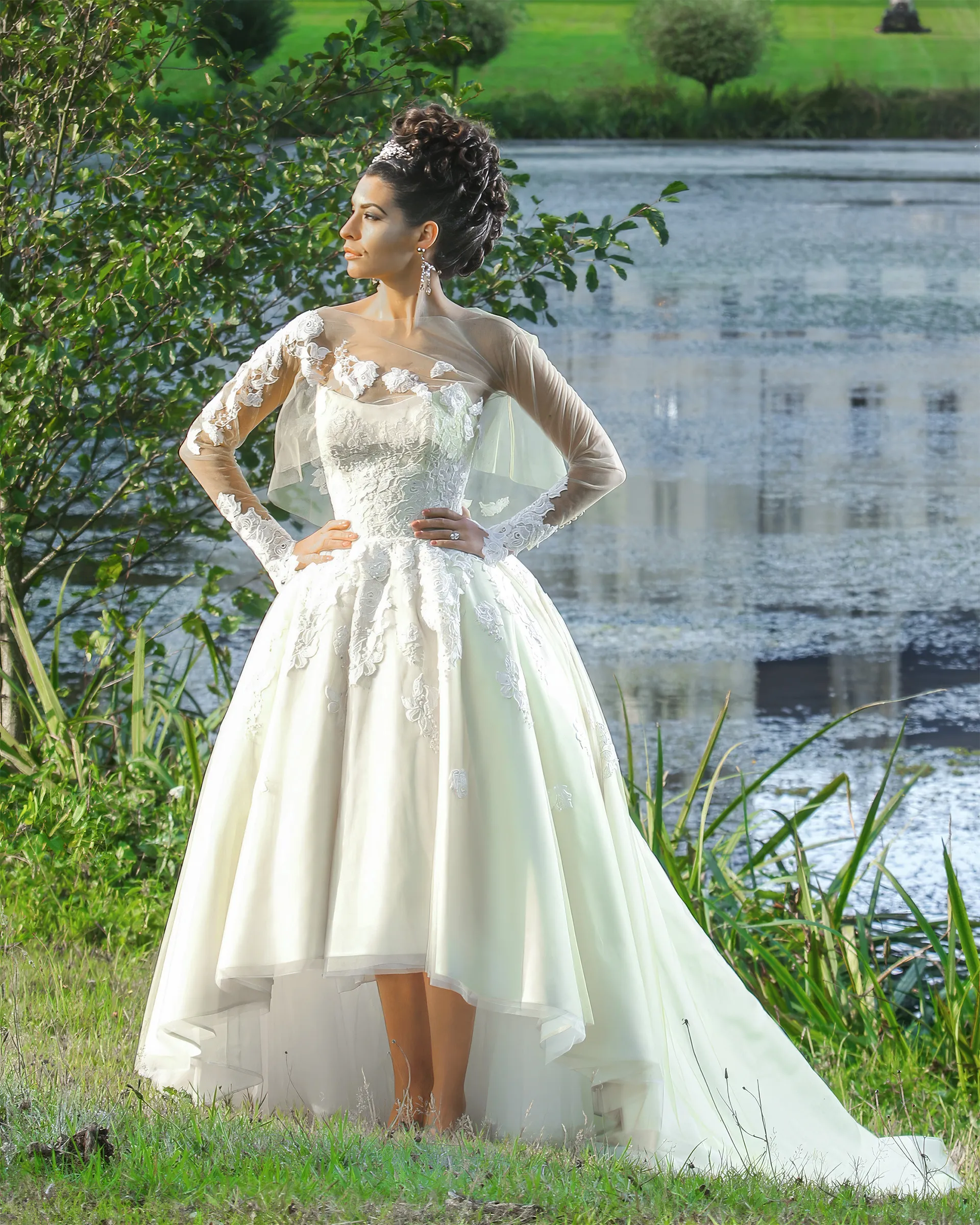 Clio by the château lake, hands on hips: guipure lace appliqués on the sheer jacket and the corseted bodice, and the skirt rising at the front to show the ankle.