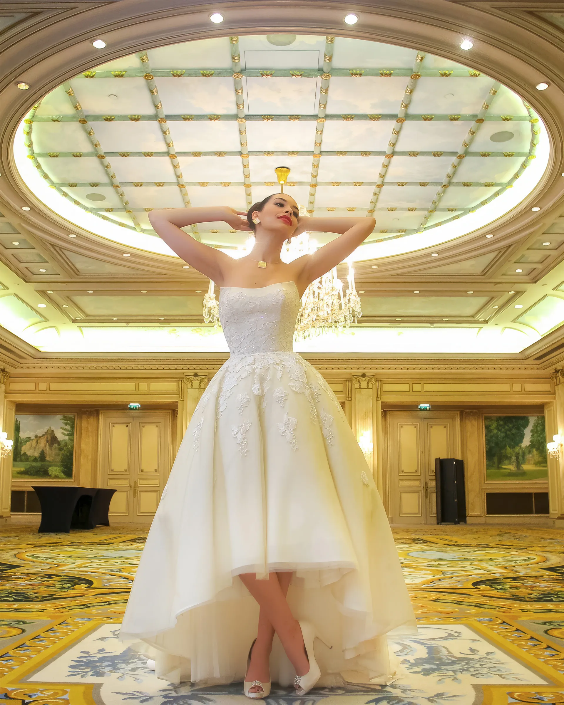 Clio without the jacket, arms raised beneath a domed ceiling in a gilded ballroom: the strapless lace bodice and the high-low tulle skirt.