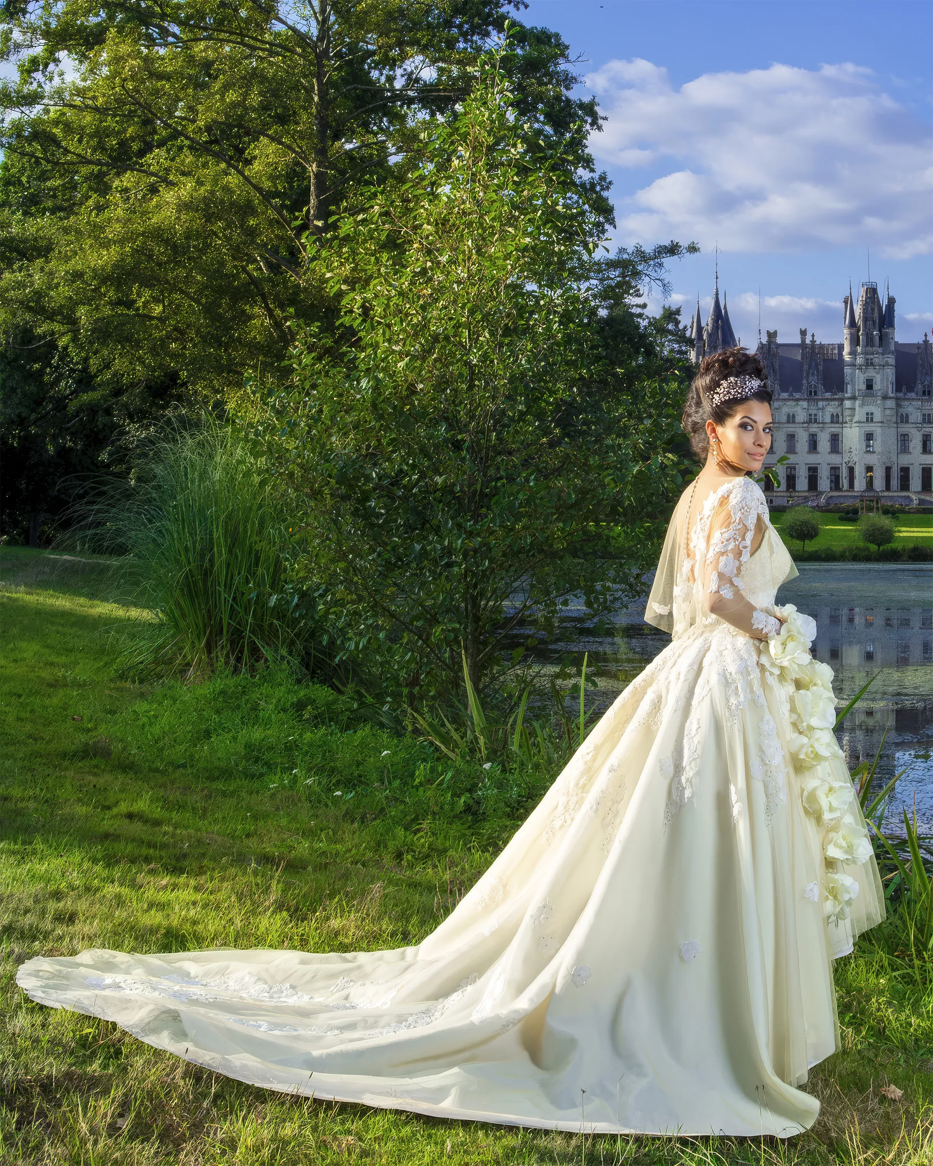 Clio in the jacket, glancing back across the château lake: lace appliqués climbing the long sleeves, a cascade of white roses and the skirt sweeping into a train.