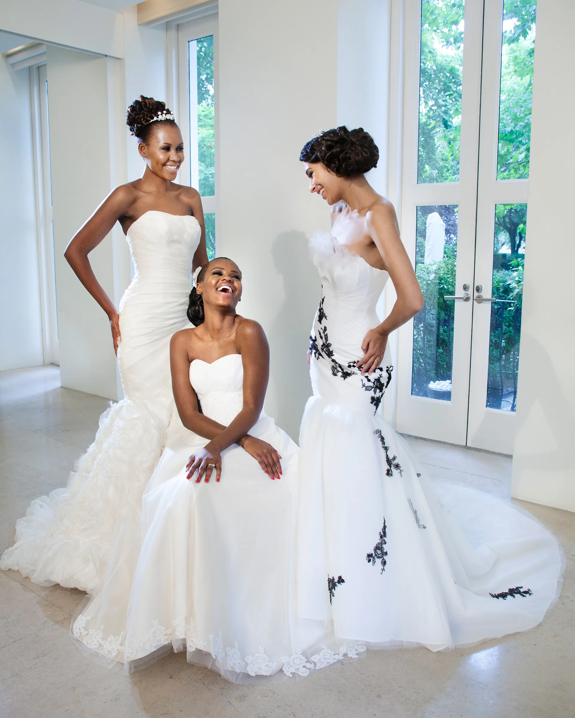 Three brides laughing together in a white room with tall garden windows: Odette's ruffled fishtail on the left, a strapless gown with a lace-edged hem in the centre, and Odile with its black lace appliqués.