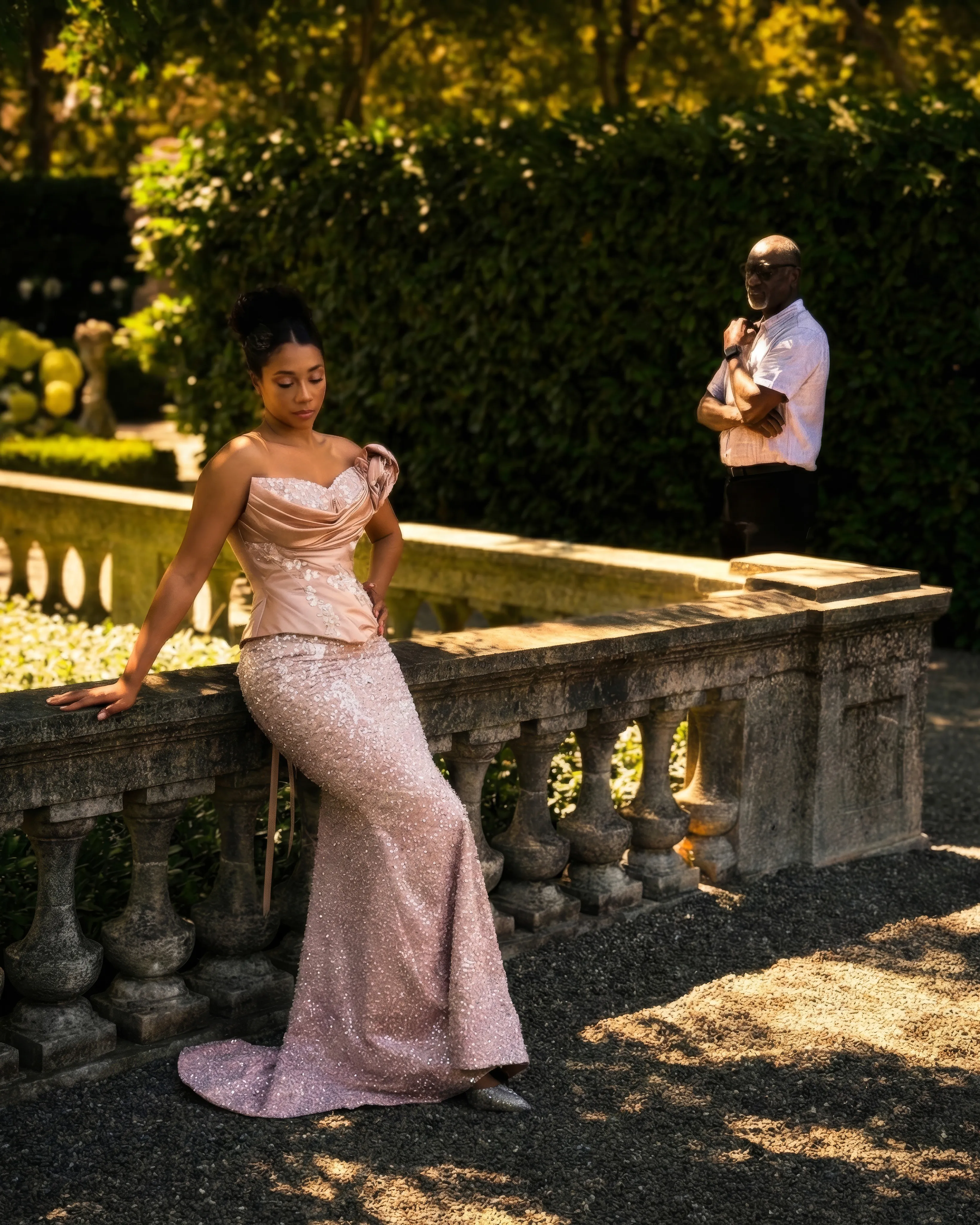 Designer and muse: Yemi Osunkoya looks on from the hedge as a model in Mist leans on a stone balustrade in a sunlit garden, the draped blush satin corset and its shoulder rose above a sequinned mermaid skirt, without its overskirt.