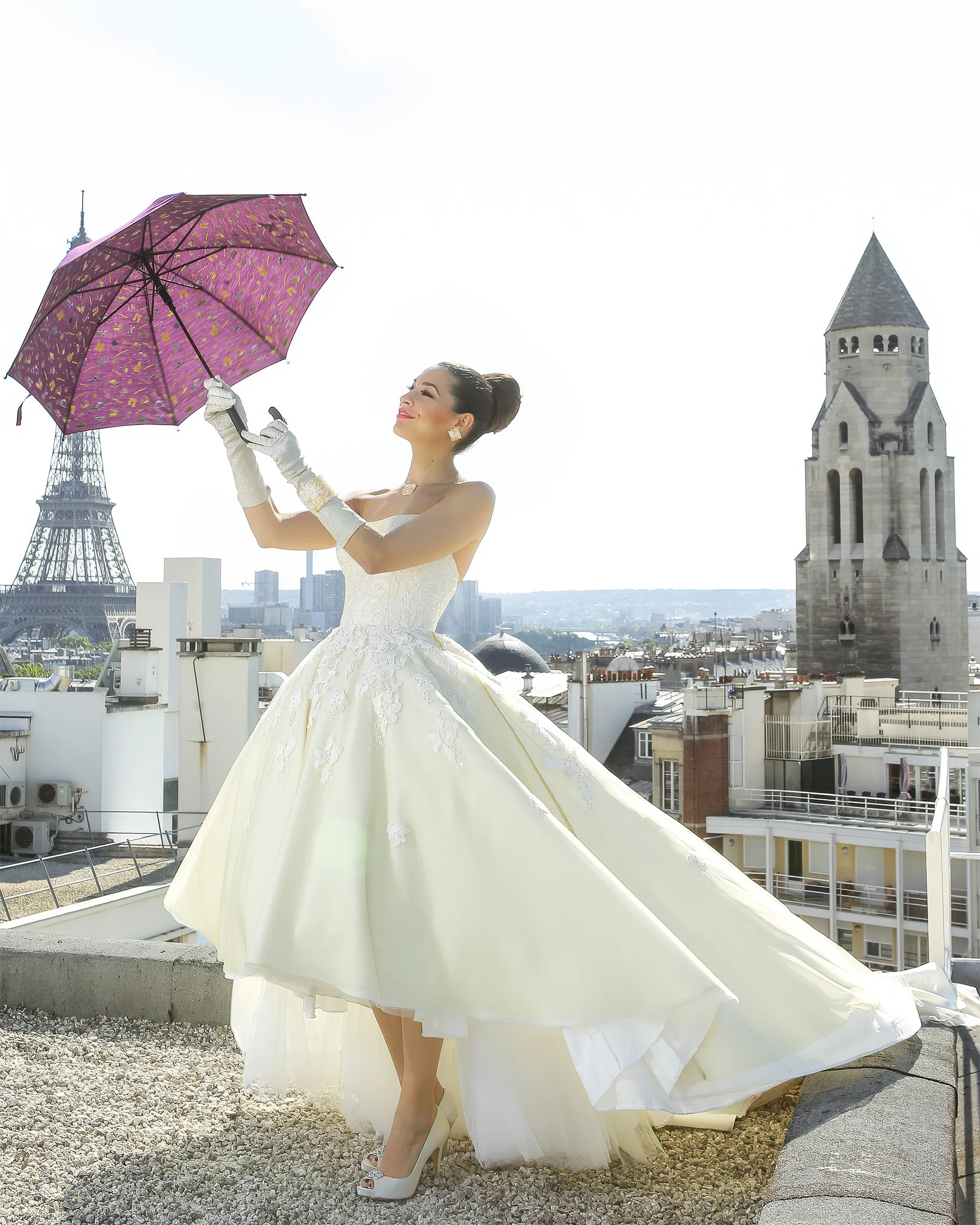 Clio on a Paris rooftop with the Eiffel Tower behind, holding a plum parasol in long white gloves: a strapless ivory gown with guipure lace appliqués and a high-low tulle skirt.